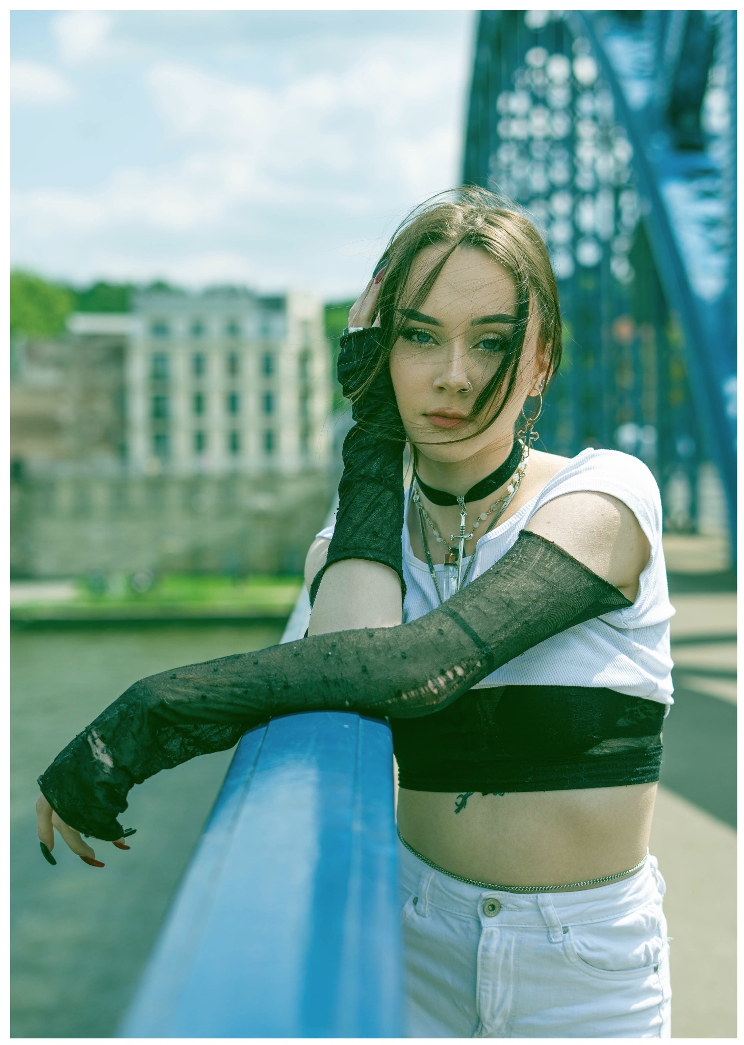 Młoda kobieta w stylizacji gotyckiej pozuje na tle niebieskiego mostu, nosząc białe spodnie, top, czarny choker i siateczkowe rękawiczki z dziurami.