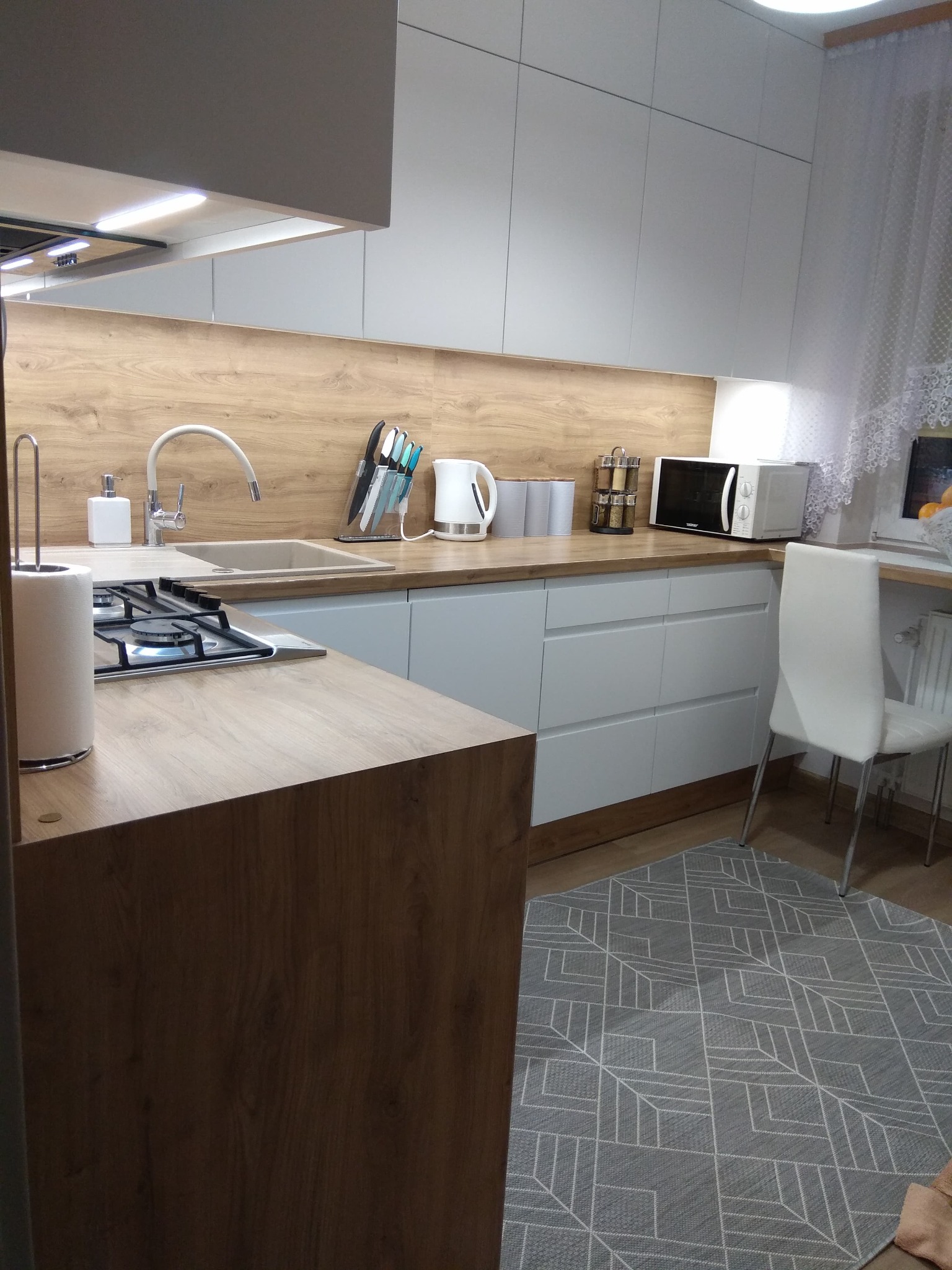 Jasna kuchnia z drewnianym blatem i białymi szafkami, widoczny narożny zlew, płyta gazowa, akcesoria kuchenne i szary dywan z geometrycznym wzorem.