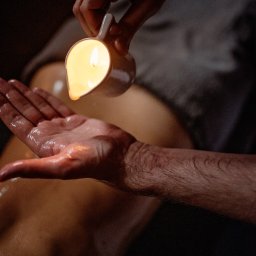 Jacek Sobolewski - Terapeuta wylewa ciepły olej z ceramicznego naczynia na dłoń klienta leżącego na stole do zabiegów, tworząc refleksy świetlne na skórze.