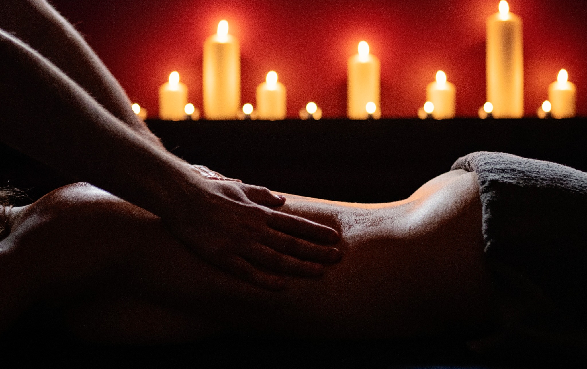 Dłonie masujące plecy kobiety w zaciemnionym pomieszczeniu, w tle rząd płonących świec na czarnej półce przed czerwoną ścianą.