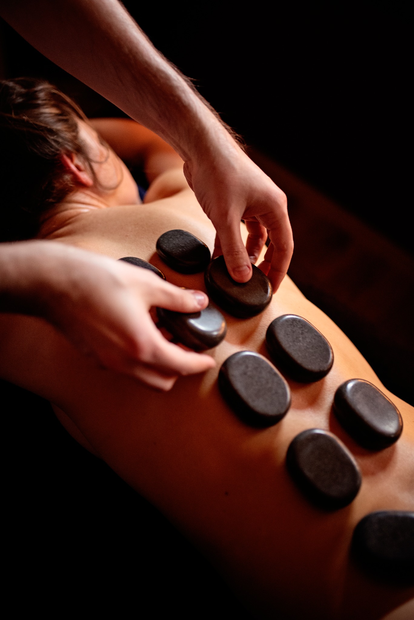 Masażysta układa rozgrzane, gładkie kamienie na plecach klienta podczas zabiegu masażu gorącymi kamieniami; widoczne dłonie terapeuty i fragment pleców pacjenta w ciepłym oświetleniu.