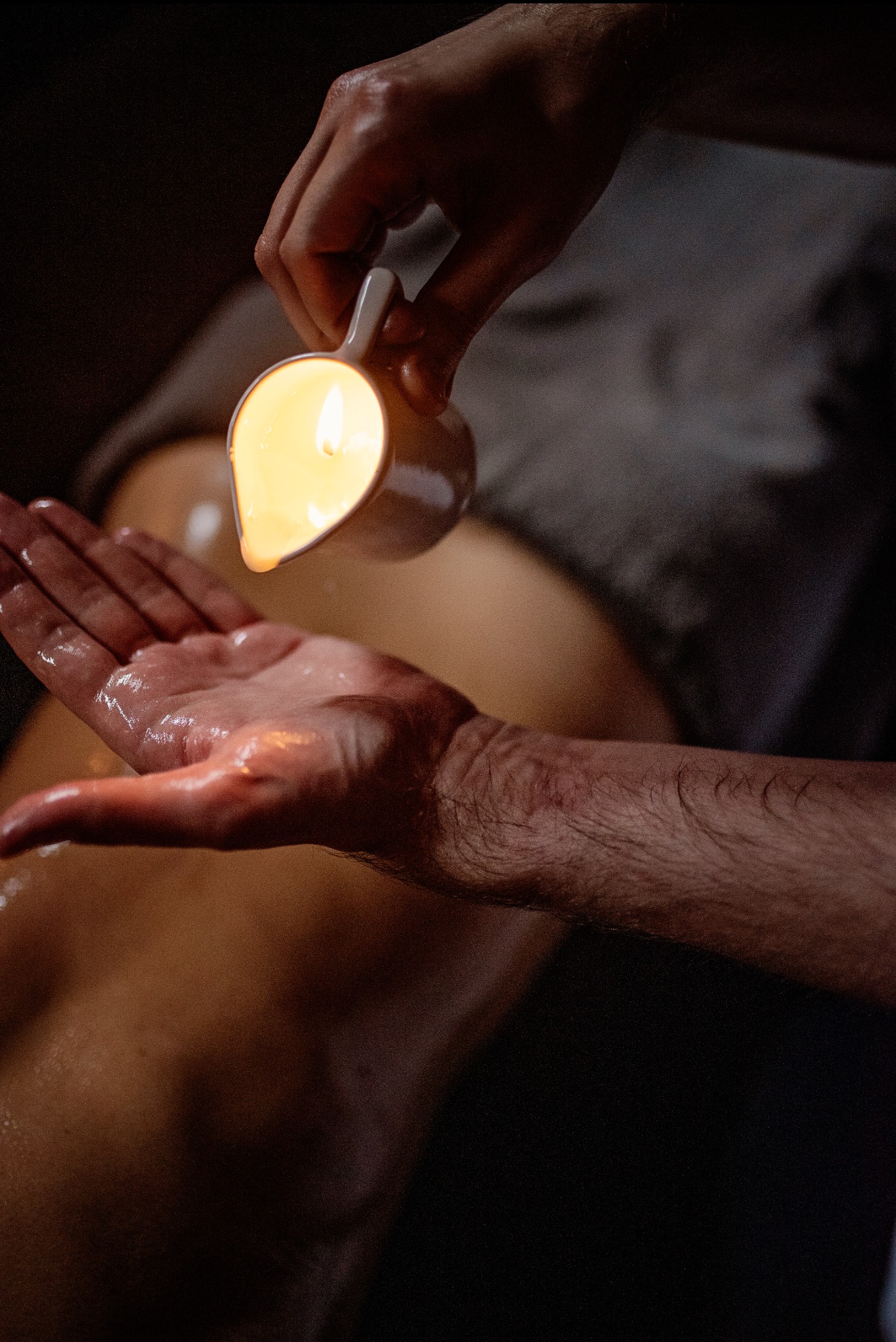 Terapeuta wylewa ciepły olej z ceramicznego naczynia na dłoń klienta leżącego na stole do zabiegów, tworząc refleksy świetlne na skórze.