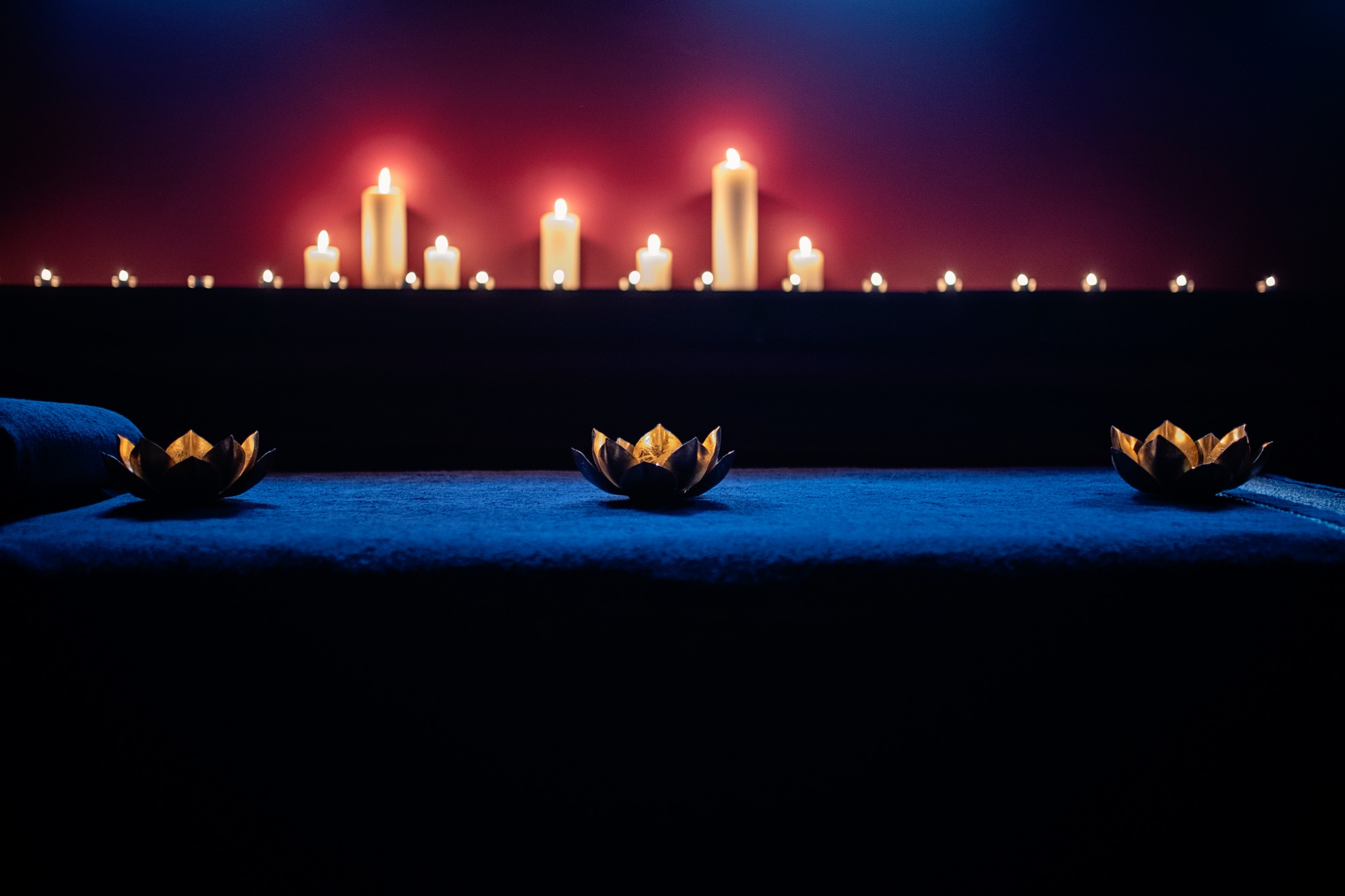 Przyciemnione wnętrze gabinetu relaksacyjnego z leżanką, na której umieszczono trzy dekoracyjne lampiony w kształcie kwiatu lotosu, w tle rząd zapalonych świec na półce podświetlonej czerwienią.