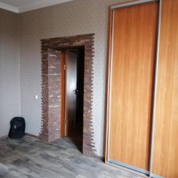 MegaBuD Viacheslav Sapko - Wykończenie wnętrza: dekoracyjne obramowanie z cegiełek wokół otwartych drzwi, drewniana podłoga i szafa przesuwna. W tle wzorzysta tapeta i biały sufit.