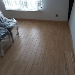 Ułożone panele podłogowe w ciepłym odcieniu drewna, widoczne listwy przypodłogowe. Pokój po remoncie w Gliwicach, meble zabezpieczone folią.