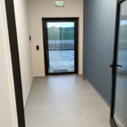 Jasne płytki podłogowe w korytarzu z drzwiami na zewnątrz, ściany białe i szaroniebieskie. Widoczny fragment ościeżnicy.