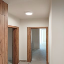 MegaBuD Viacheslav Sapko - Jasne wnętrze z drewnianą zabudową i ościeżnicami. Widoczne dwa otwory drzwiowe i fragment szafy. Na suficie okrągła lampa. Minimalistyczny design.