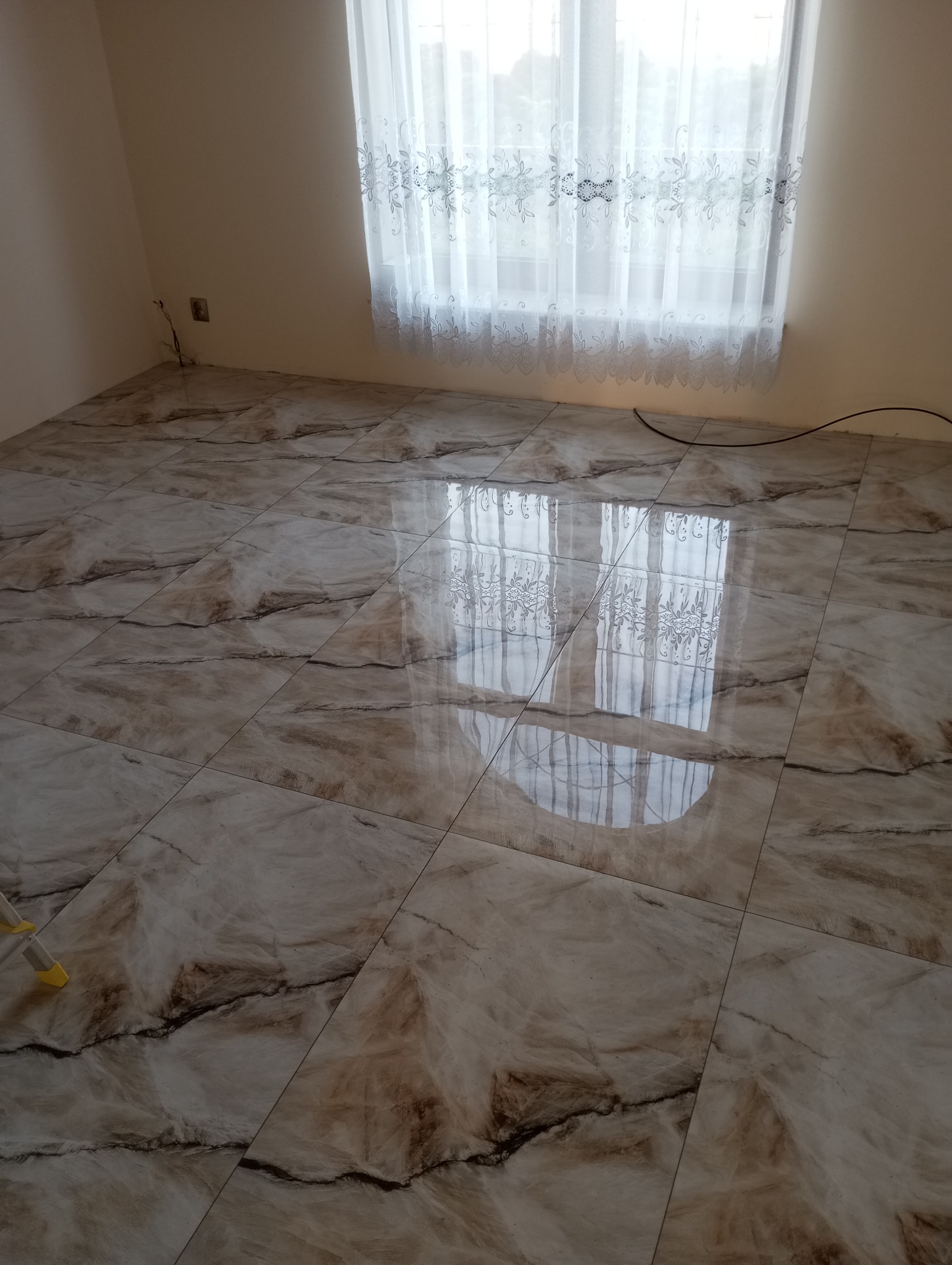 Połyskująca podłoga z płytek imitujących marmur w pokoju z oknem przesłoniętym białą, wzorzystą firanką. Widoczne odbicie okna w polerowanej powierzchni.