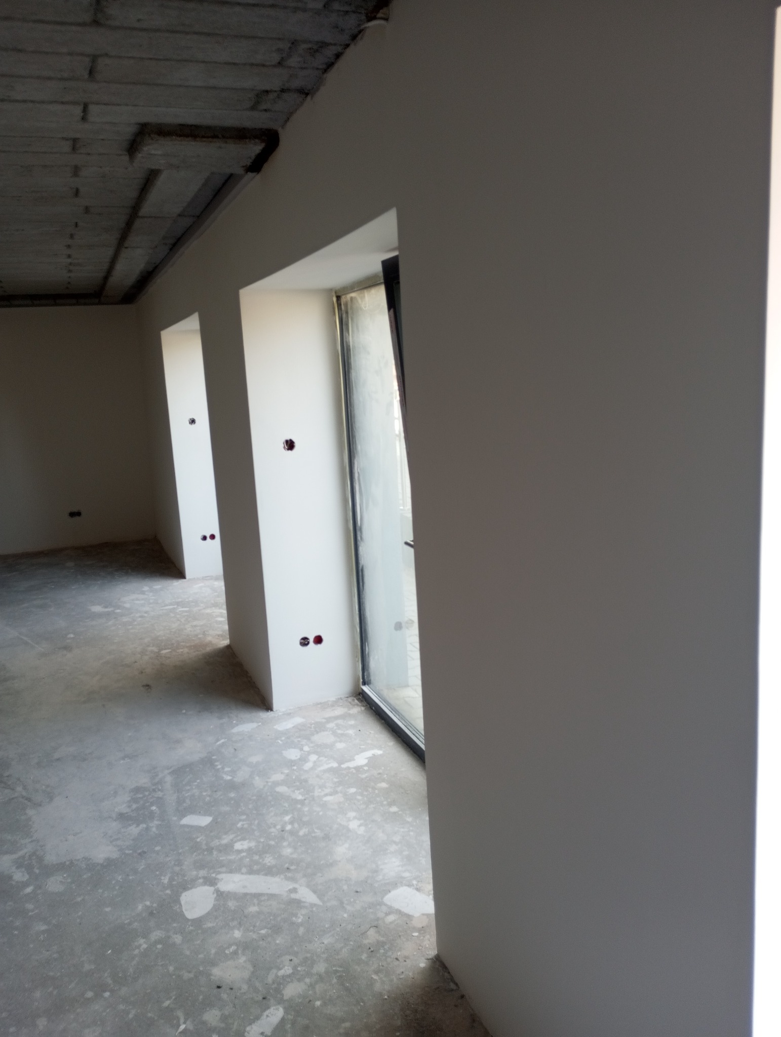 Wnętrze w trakcie wykańczania: gładkie, pomalowane ściany z otworami na gniazdka, surowy betonowy strop i surowa podłoga. Widoczne okna.