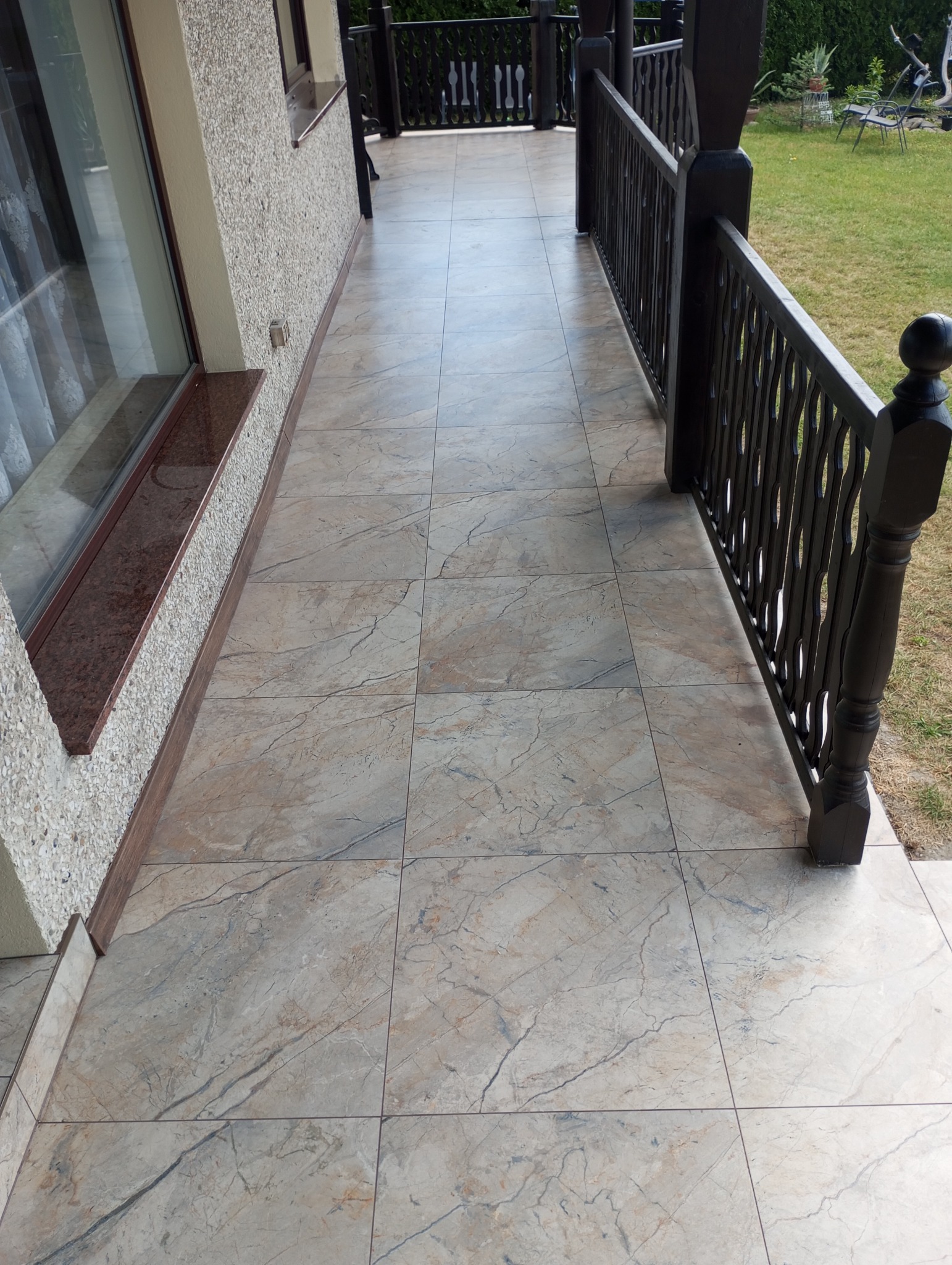 Wykończony taras z marmurowymi płytkami i ciemną, drewnianą balustradą. Widok na trawnik w tle. Estetyczne połączenie materiałów i kolorów.