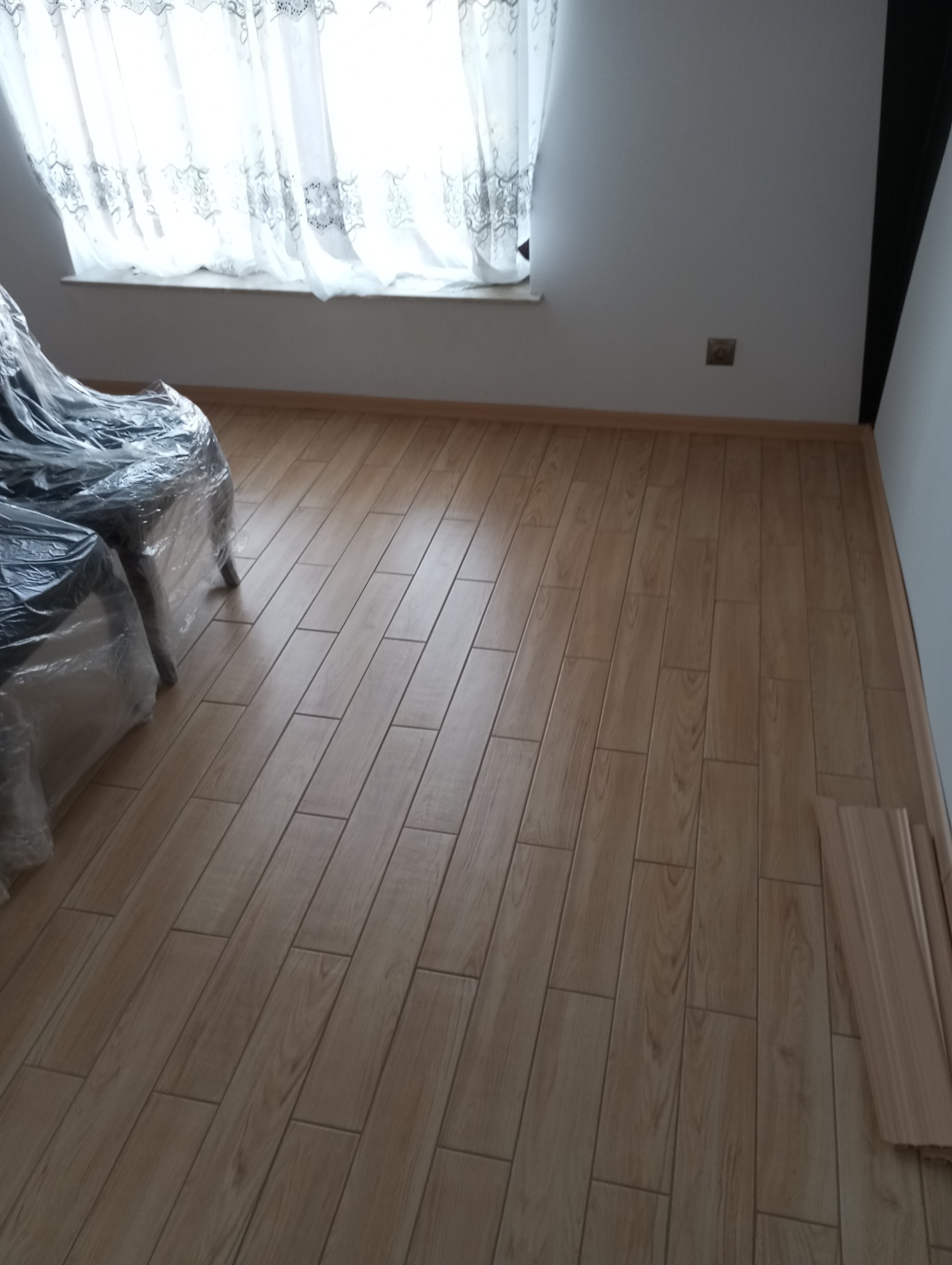 Ułożone panele podłogowe w ciepłym odcieniu drewna, widoczne listwy przypodłogowe. Pokój po remoncie w Gliwicach, meble zabezpieczone folią.