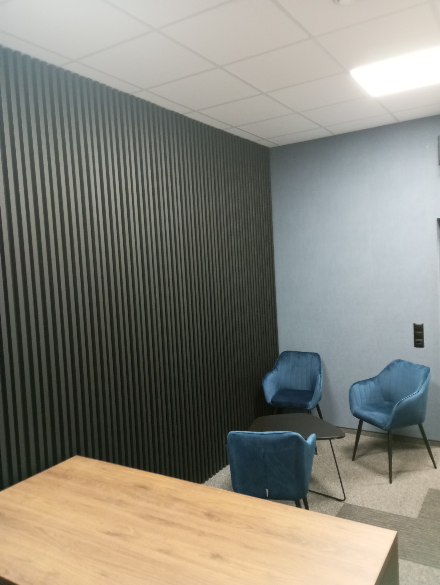Nowoczesne wykończenie biura: lamele dekoracyjne w kolorze grafitowym, błękitne krzesła i drewniany blat biurka. Stonowana kolorystyka i minimalizm.