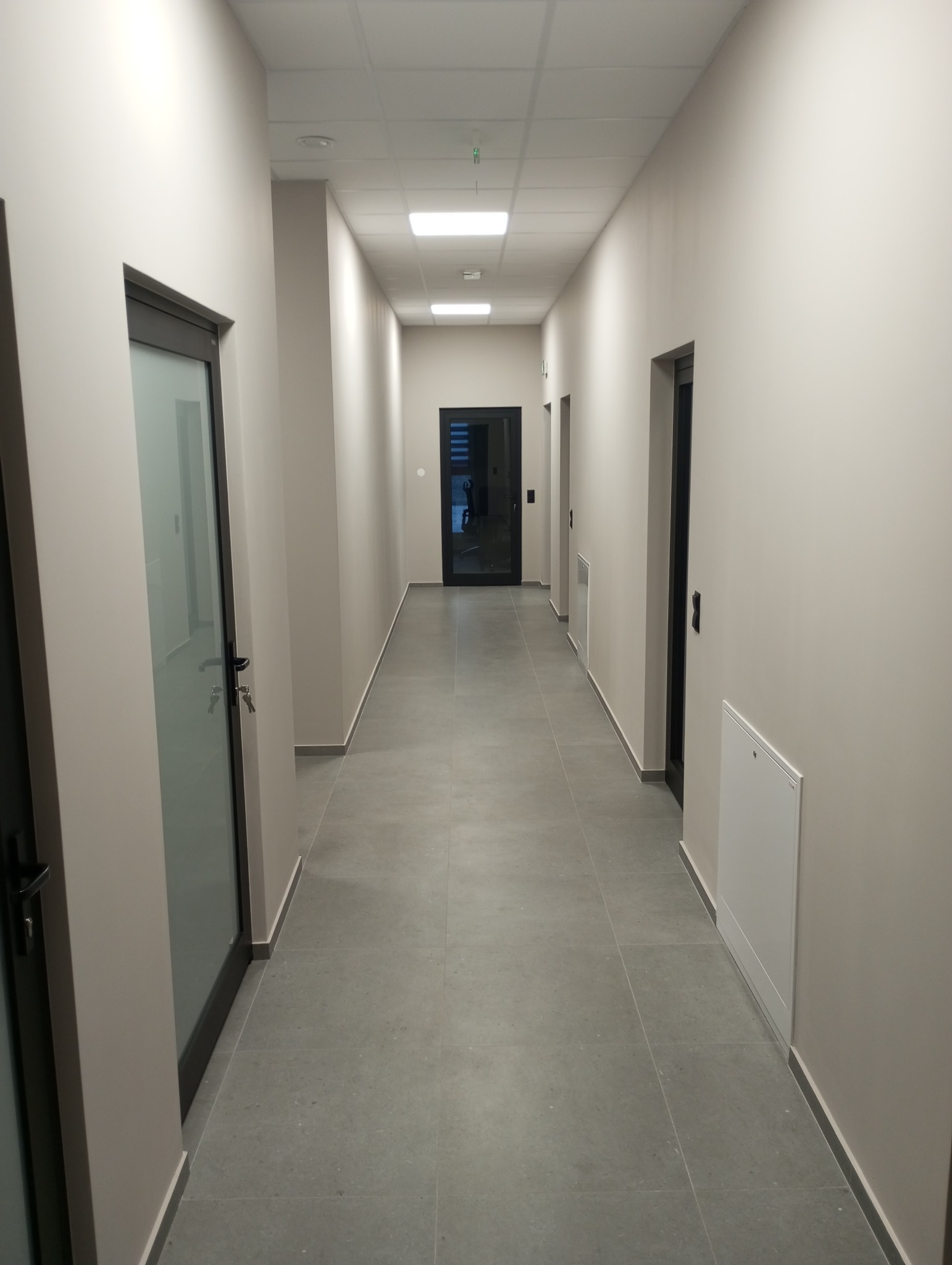 Korytarz biurowy po remoncie: szare płytki podłogowe, świeżo pomalowane ściany w odcieniach beżu, czarne ramy drzwi. Widok na wprost, oświetlenie sufitowe.