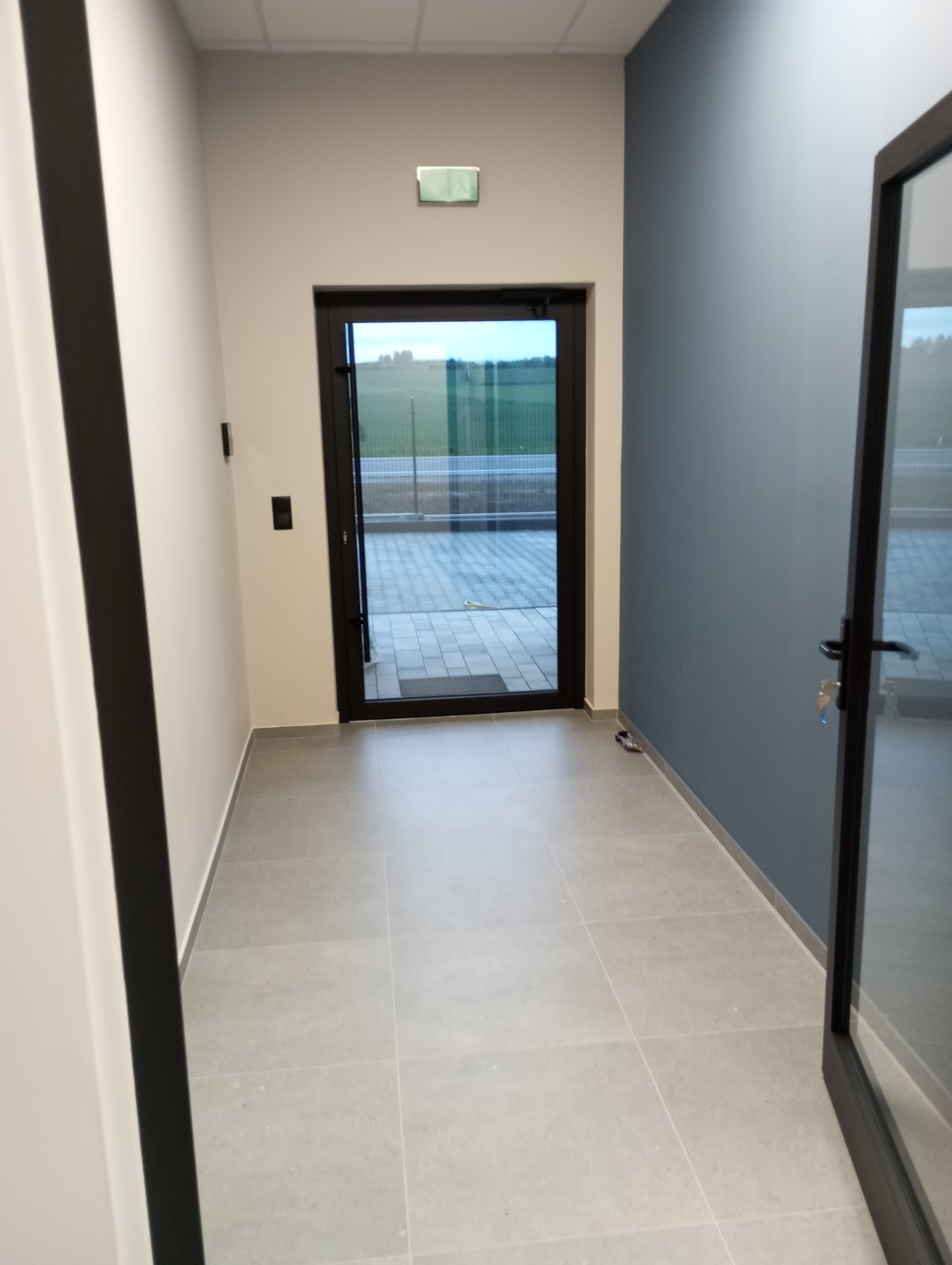 Jasne płytki podłogowe w korytarzu z drzwiami na zewnątrz, ściany białe i szaroniebieskie. Widoczny fragment ościeżnicy.