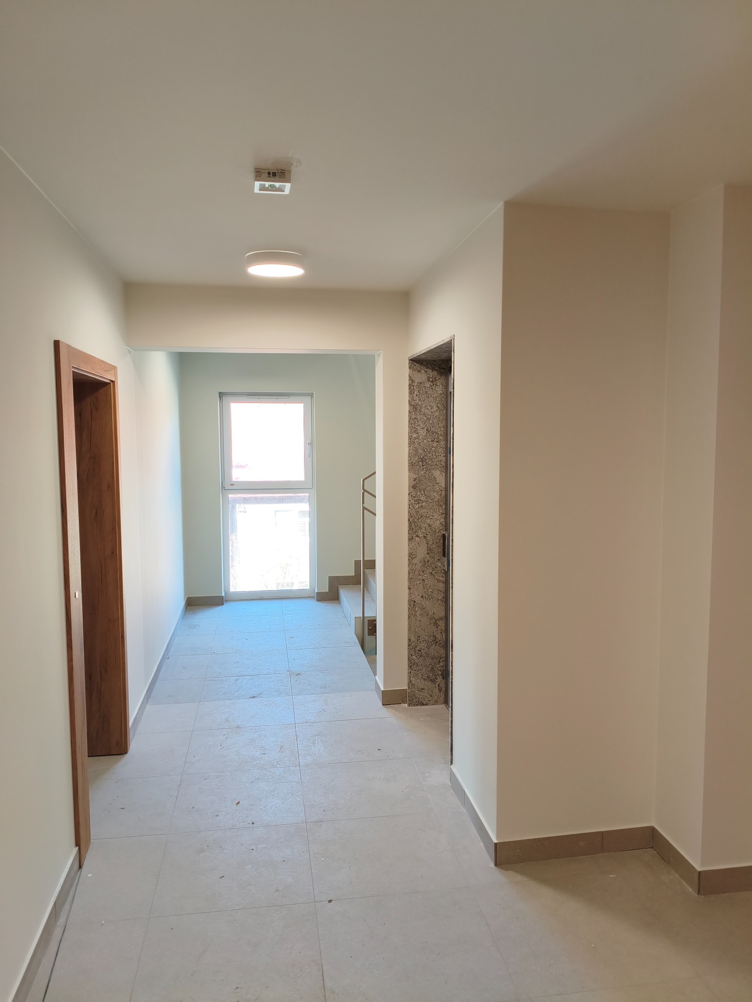 Jasny korytarz z beżowymi ścianami i szarą podłogą z płytek. Widoczne drzwi z drewnianą ościeżnicą i okno z widokiem na schody. Minimalistyczne oświetlenie sufitowe.