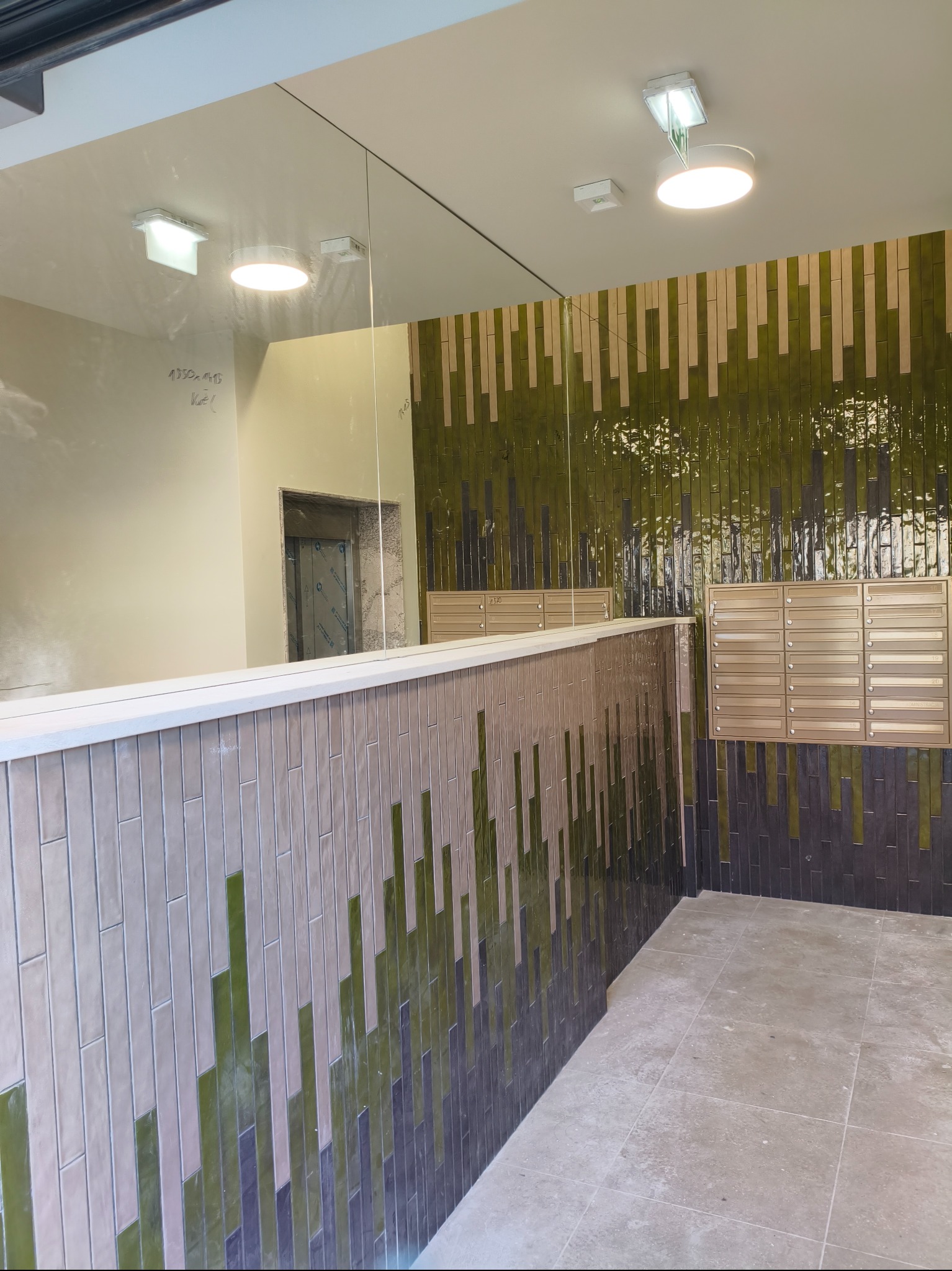 Korytarz z dekoracyjną okładziną ścienną z płytek w odcieniach zieleni i beżu, skrzynki pocztowe w kolorze złotym, duże lustro na ścianie. Nowoczesny design.