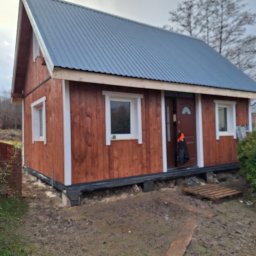 Korektor - Niewielki, brązowy dom szkieletowy z szarą, metalową blachodachówką, białymi obramowaniami okien i drzwiami wejściowymi, stojący na fundamencie, z widocznym błotnistym terenem i paletami przed...