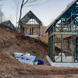 Korektor - Dwa domy w trakcie budowy, z widoczną konstrukcją szkieletową, jeden z nich pomalowany na zielono, na nierównym terenie z widocznymi materiałami budowlanymi i zbrojeniem.