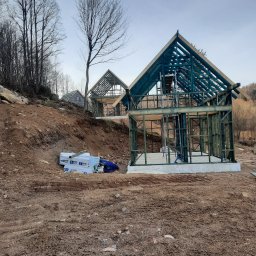 Korektor - Wznoszenie konstrukcji z drewna na zboczu wzgórza, widoczne fundamenty i materiały budowlane, w tle drzewa bez liści i zalesione wzgórze.