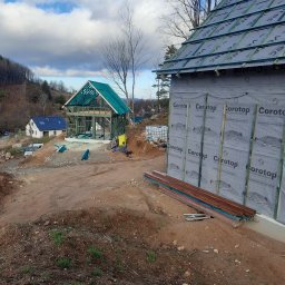 Korektor - Wznoszenie konstrukcji dwóch budynków w technologii szkieletowej, jeden w stanie surowym otwartym z widoczną ramą, drugi z częściowo wykończonym dachem i ścianami zewnętrznymi pokrytymi membraną.