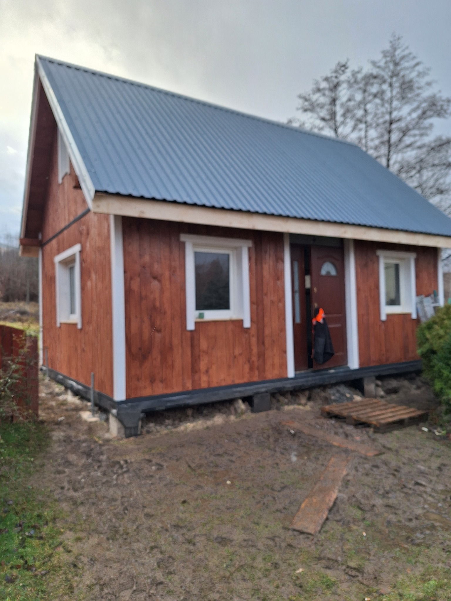 Niewielki, brązowy dom szkieletowy z szarą, metalową blachodachówką, białymi obramowaniami okien i drzwiami wejściowymi, stojący na fundamencie, z widocznym błotnistym terenem i paletami przed...