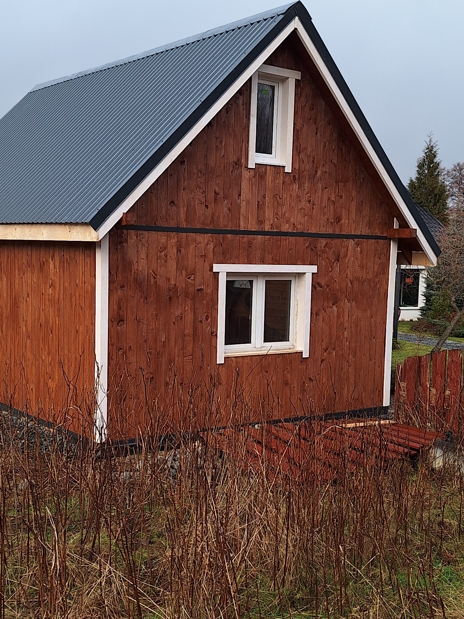 Brunatny budynek z drewnianą elewacją, szarą blachą falistą na dachu i białymi obramowaniami okien, widziany z perspektywy niskiej trawy.