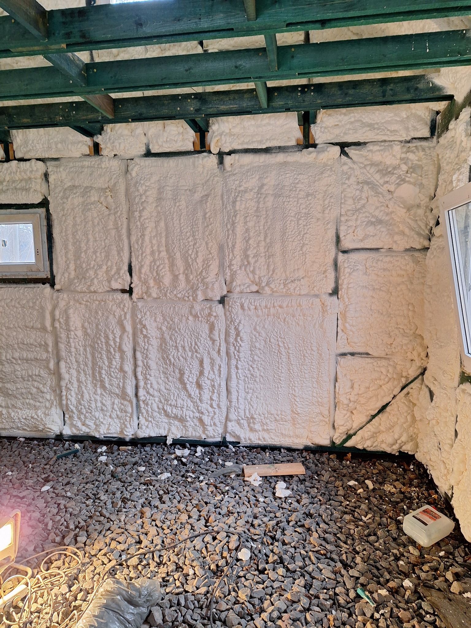 Wnętrze w trakcie izolacji pianą PUR, widoczne drewniane elementy konstrukcji, okno oraz gruz na podłodze.