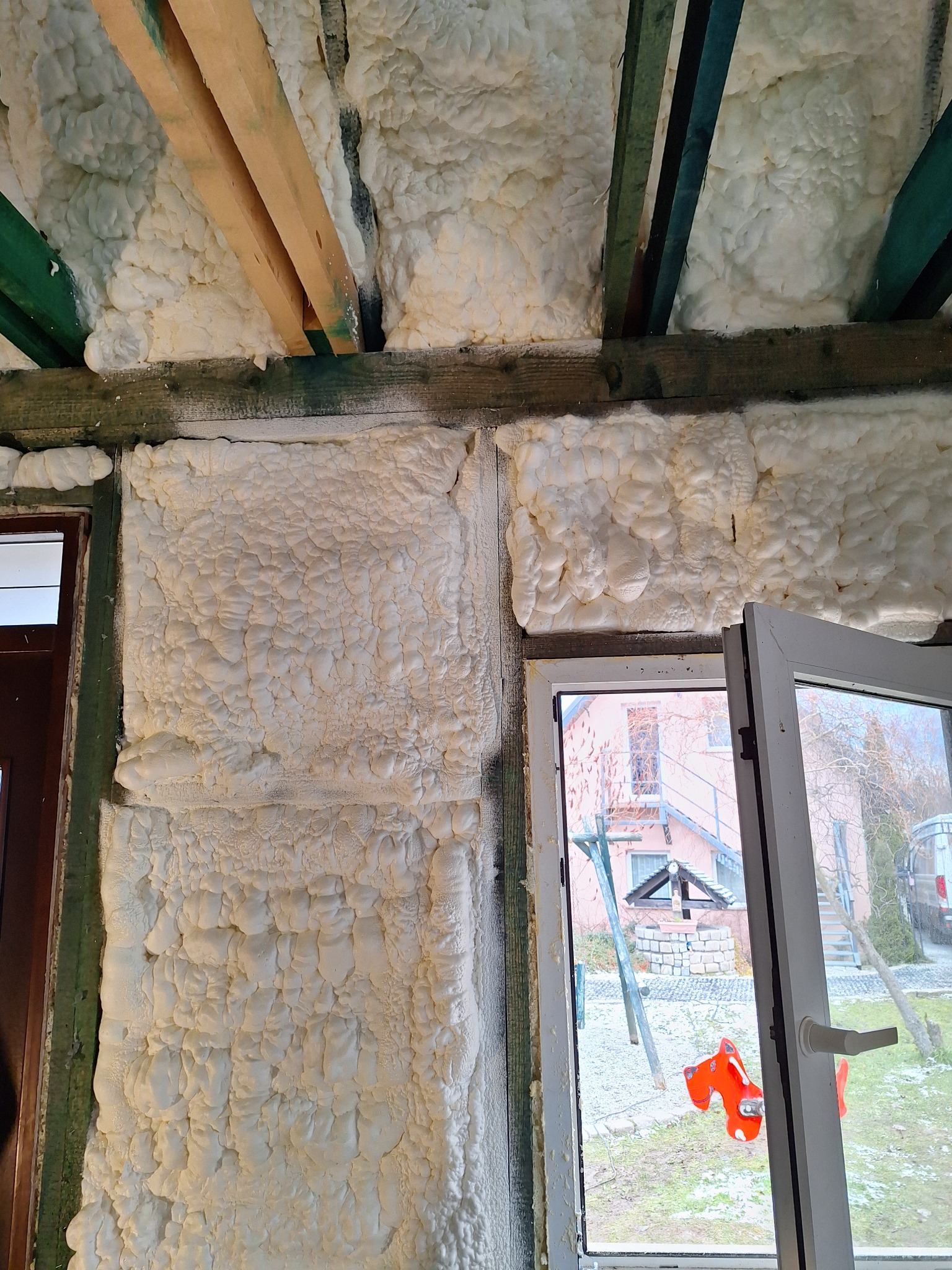 Izolacja pianką poliuretanową na ścianach i suficie budynku z drewnianą konstrukcją, widok na zewnątrz przez otwarte okno z widocznym placem zabaw i studnią.