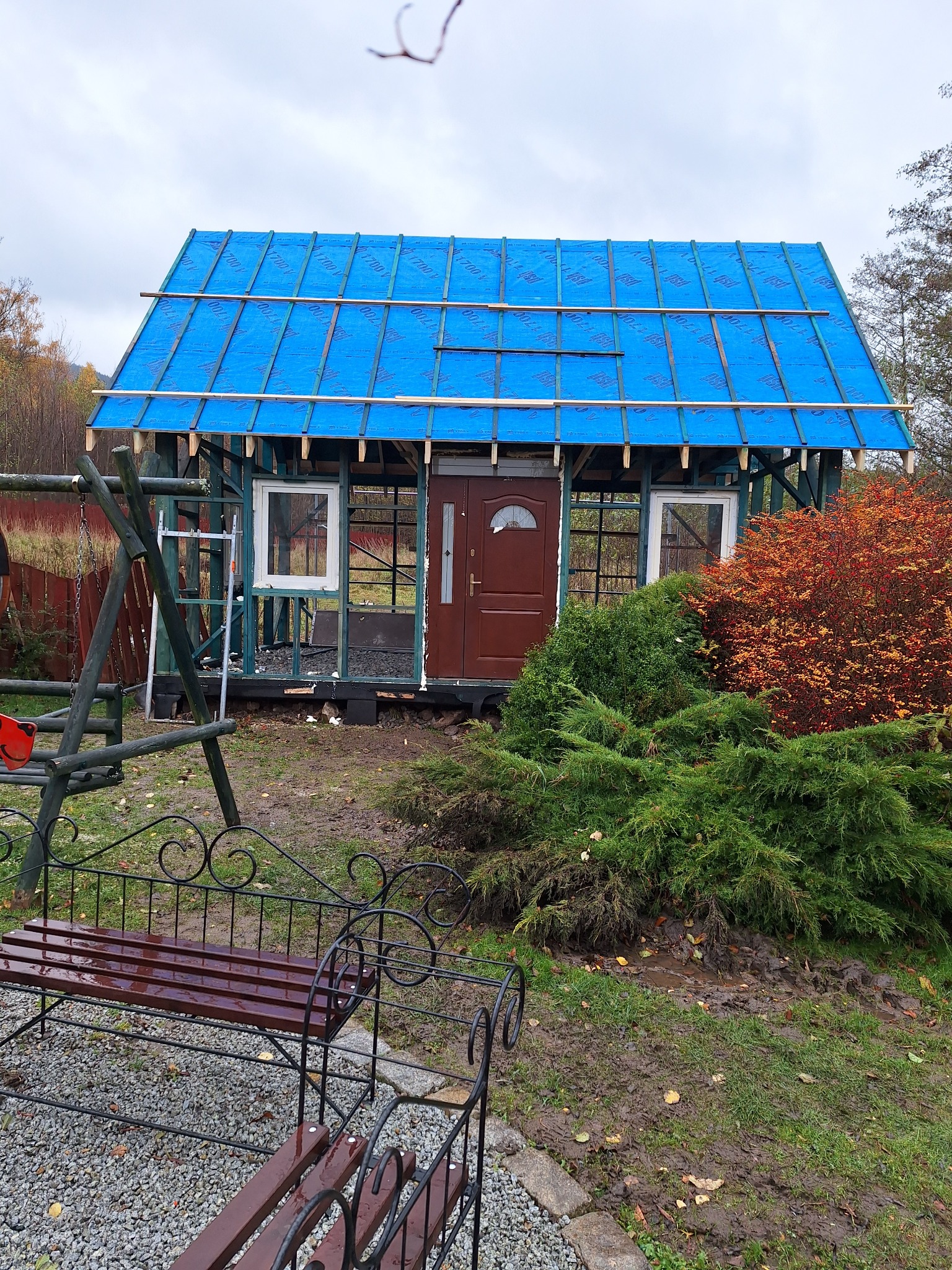 Niedokończona konstrukcja budynku z drewnianym szkieletem i niebieską folią na dachu, widoczna drabina i fragment huśtawki na zewnątrz, obok rosną krzewy.