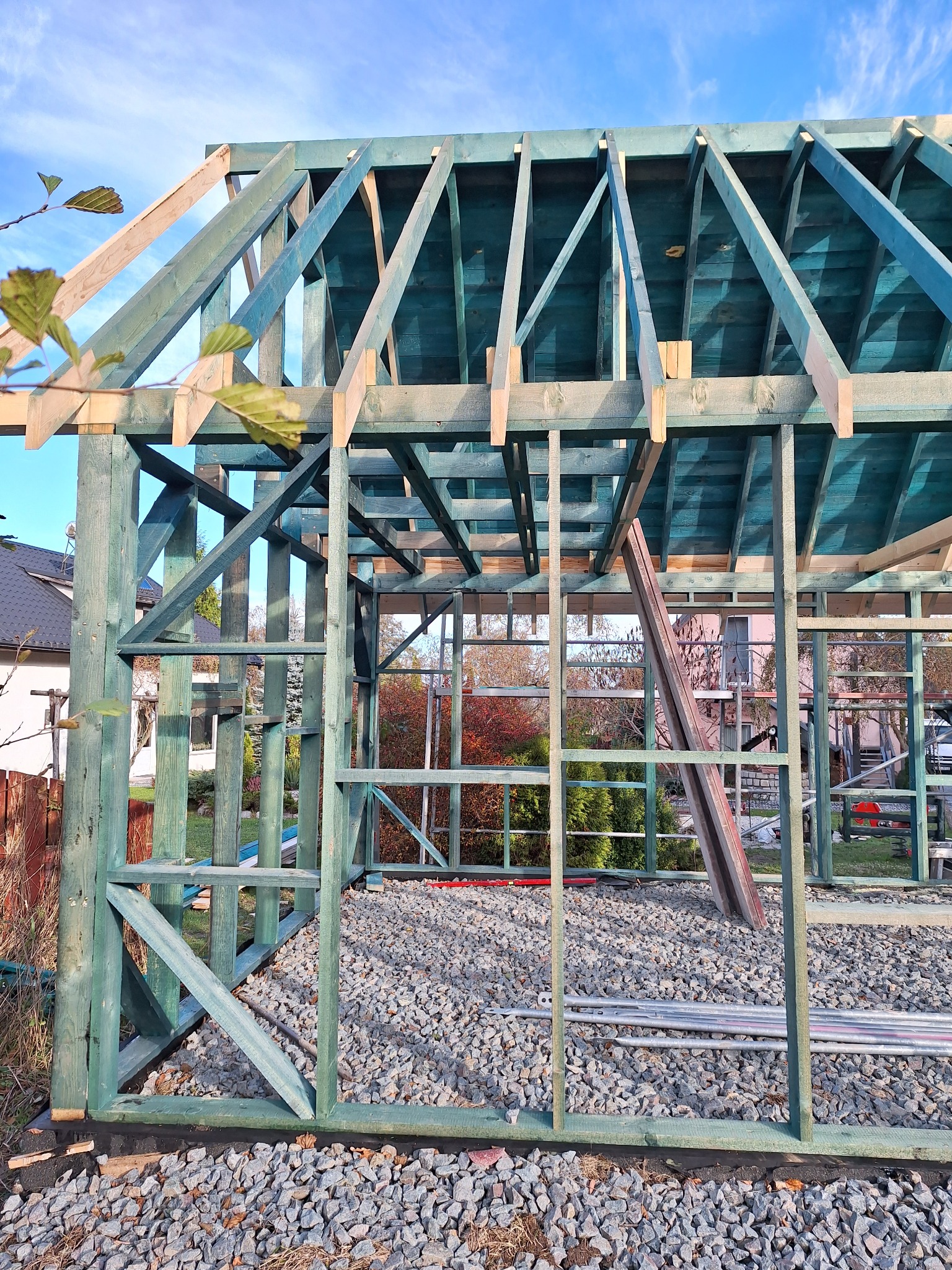 Konstrukcja szkieletowa w kolorze morskim, widoczne elementy dachu i ścian, otoczona żwirem i rurami, na tle zieleni ogrodu i błękitnego nieba.