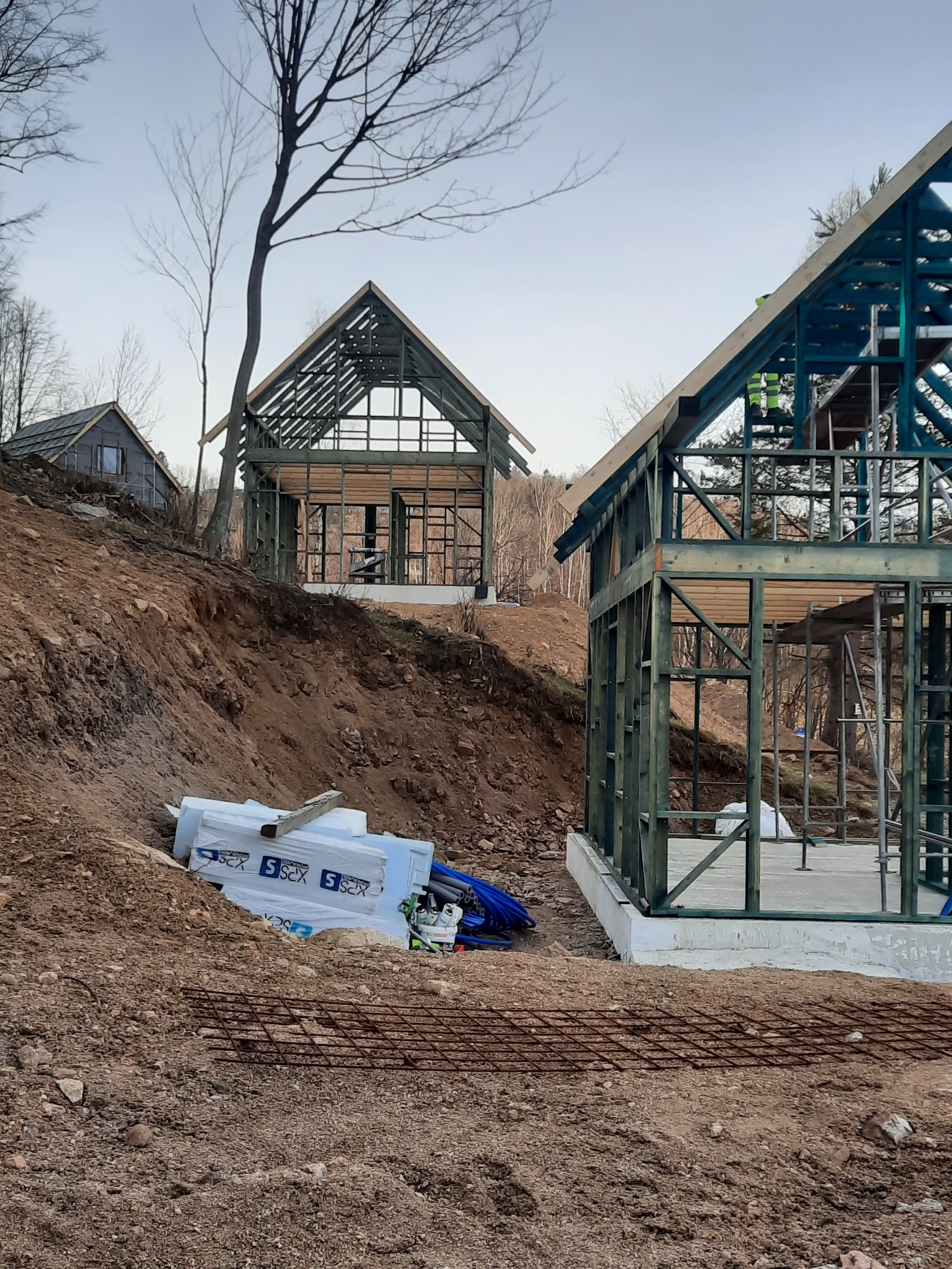 Dwa domy w trakcie budowy, z widoczną konstrukcją szkieletową, jeden z nich pomalowany na zielono, na nierównym terenie z widocznymi materiałami budowlanymi i zbrojeniem.