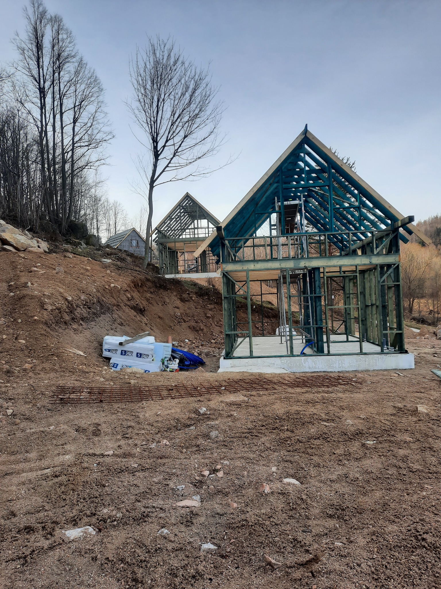 Wznoszenie konstrukcji z drewna na zboczu wzgórza, widoczne fundamenty i materiały budowlane, w tle drzewa bez liści i zalesione wzgórze.