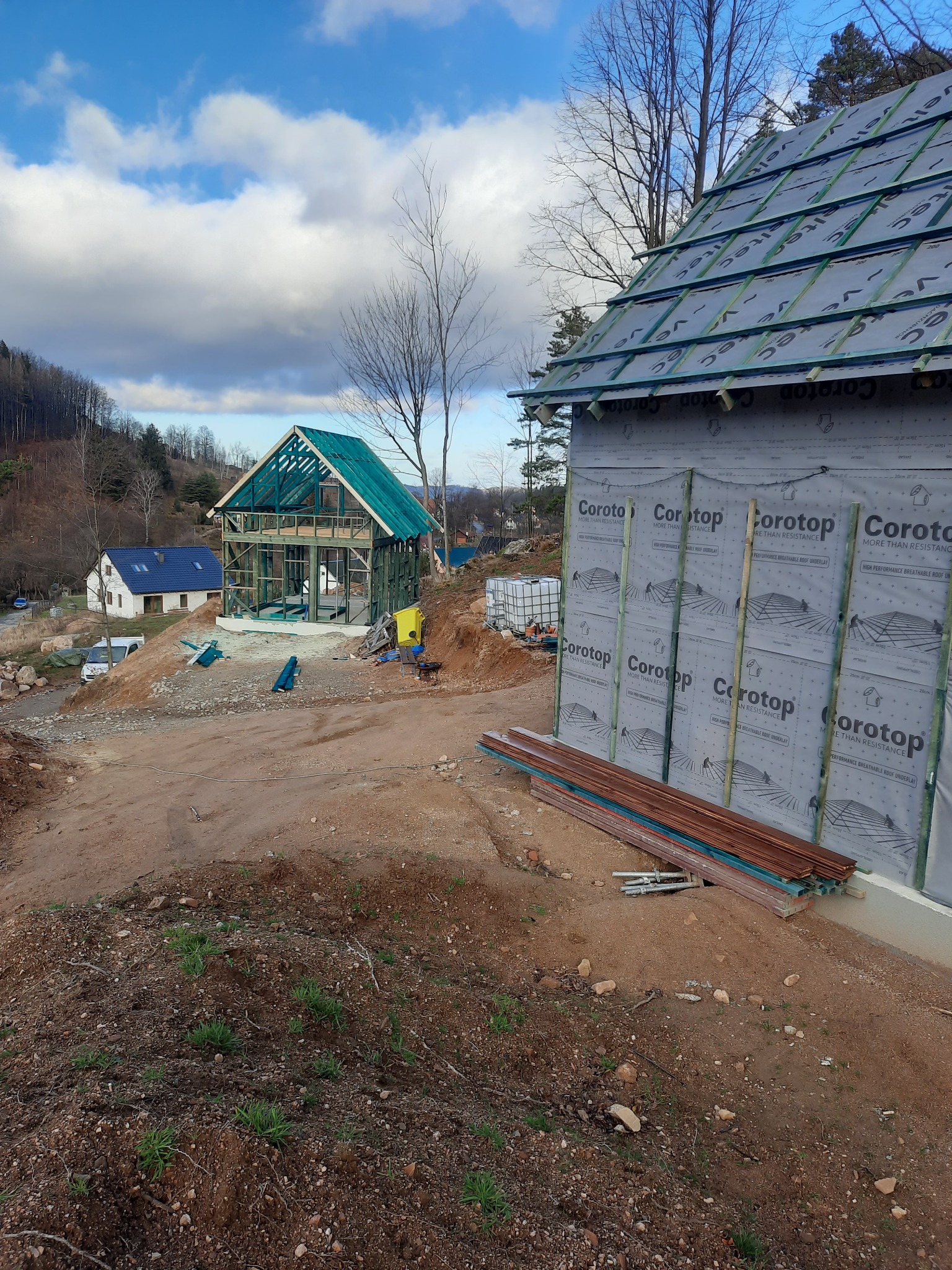 Wznoszenie konstrukcji dwóch budynków w technologii szkieletowej, jeden w stanie surowym otwartym z widoczną ramą, drugi z częściowo wykończonym dachem i ścianami zewnętrznymi pokrytymi membraną.