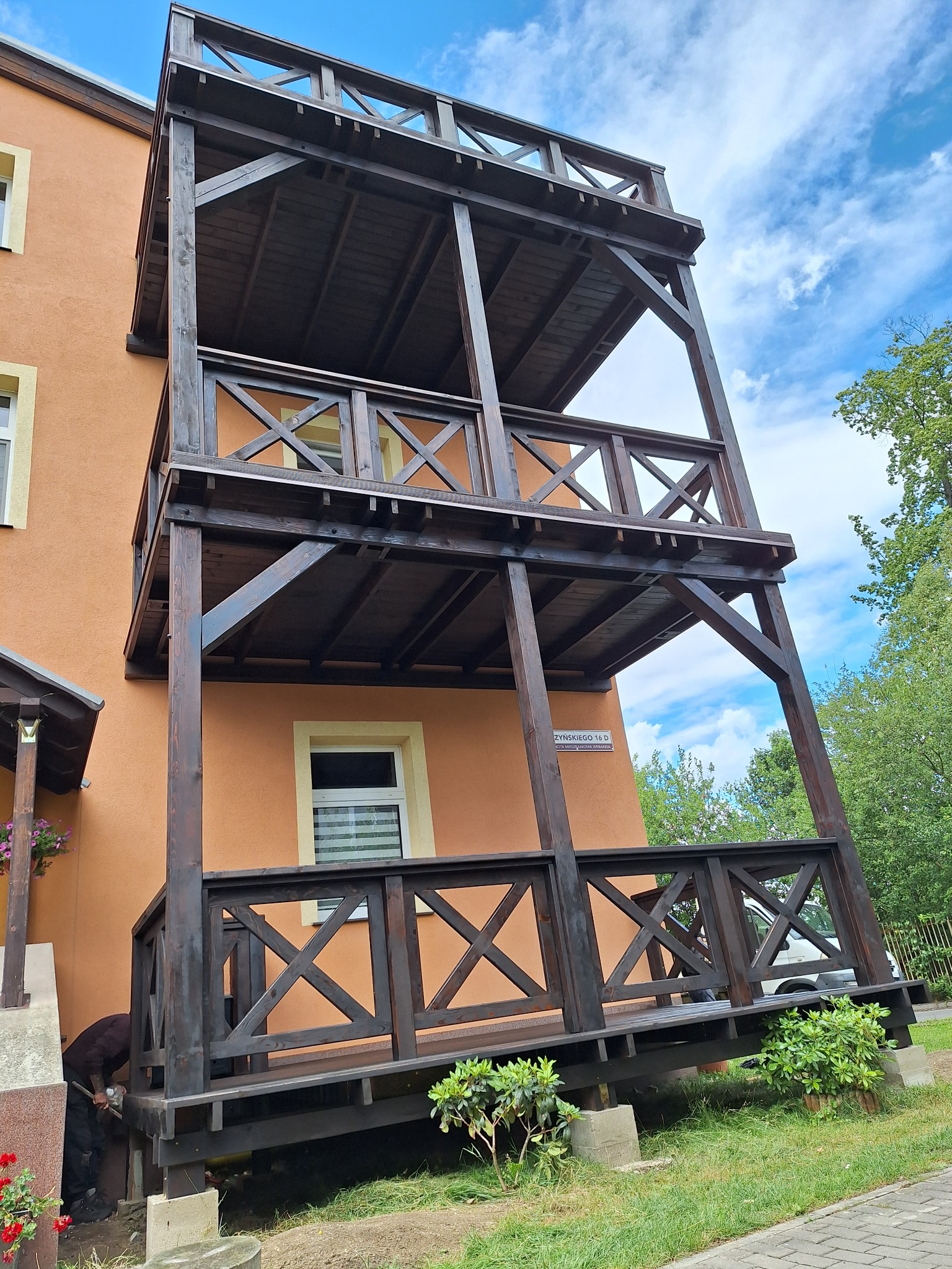 Dwukondygnacyjny taras drewniany z balustradami krzyżowymi, przylegający do pomarańczowego budynku, widok z dołu, częściowo zasłonięty przez pracującą osobę.