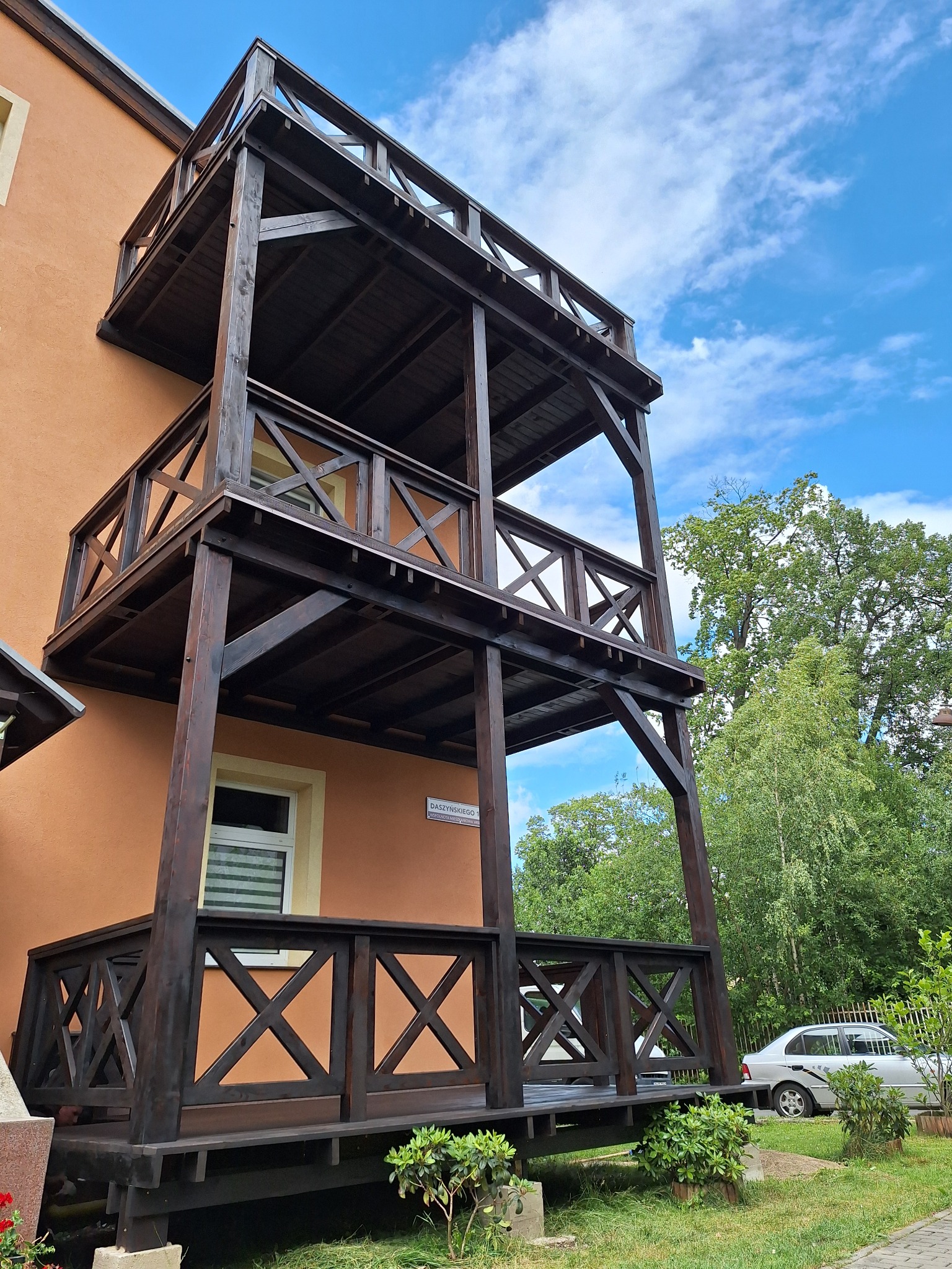 Trzykondygnacyjny, ciemnobrązowy taras drewniany przylegający do pomarańczowego budynku, widoczny z dołu, z błękitnym niebem i drzewami w tle.