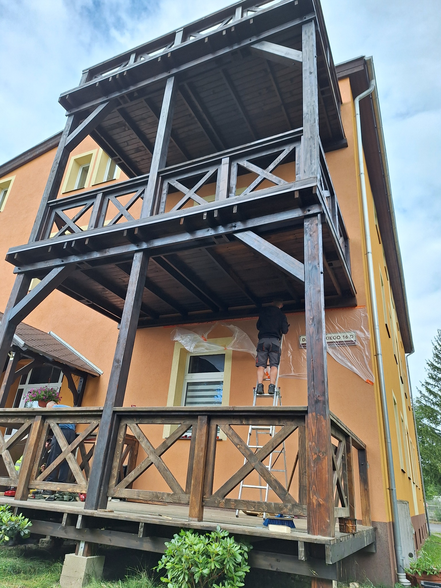 Malowanie drewnianej konstrukcji dwupoziomowego tarasu przy budynku. Mężczyzna na drabinie zabezpiecza ścianę folią. Widoczne elementy tarasu z krzyżowymi zdobieniami.
