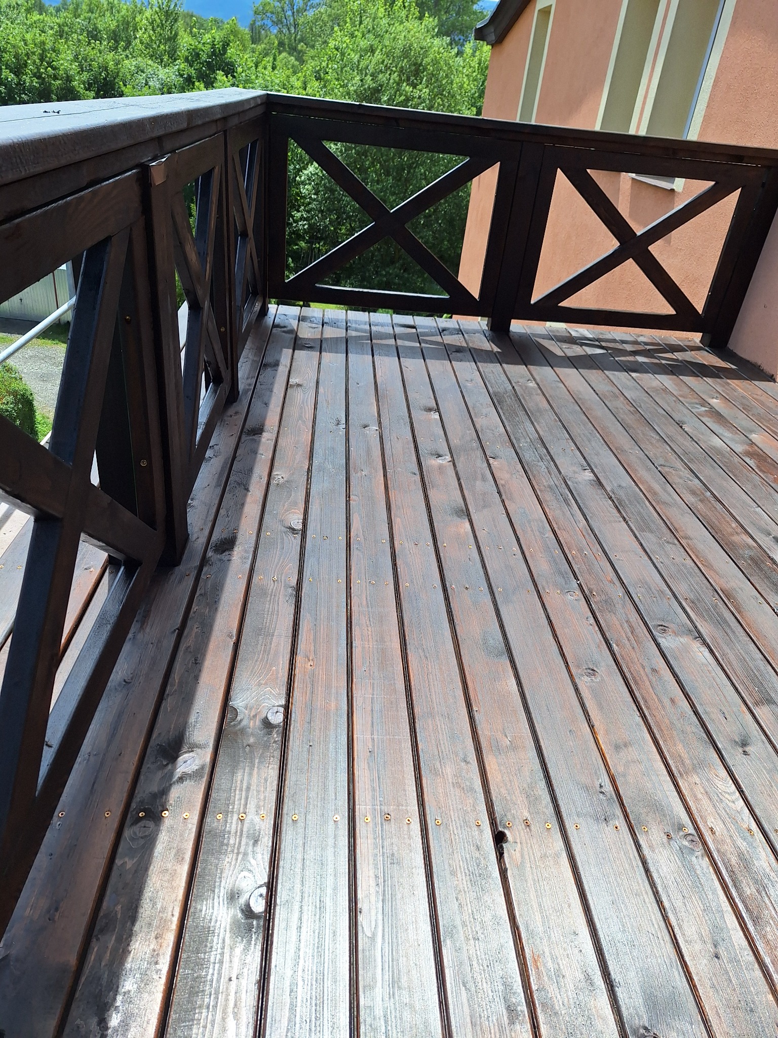 Drewniany taras z ciemnobrązową balustradą o geometrycznym wzorze krzyży, widok z góry na deski z widocznymi śrubami, w tle zieleń drzew i fragment elewacji budynku.