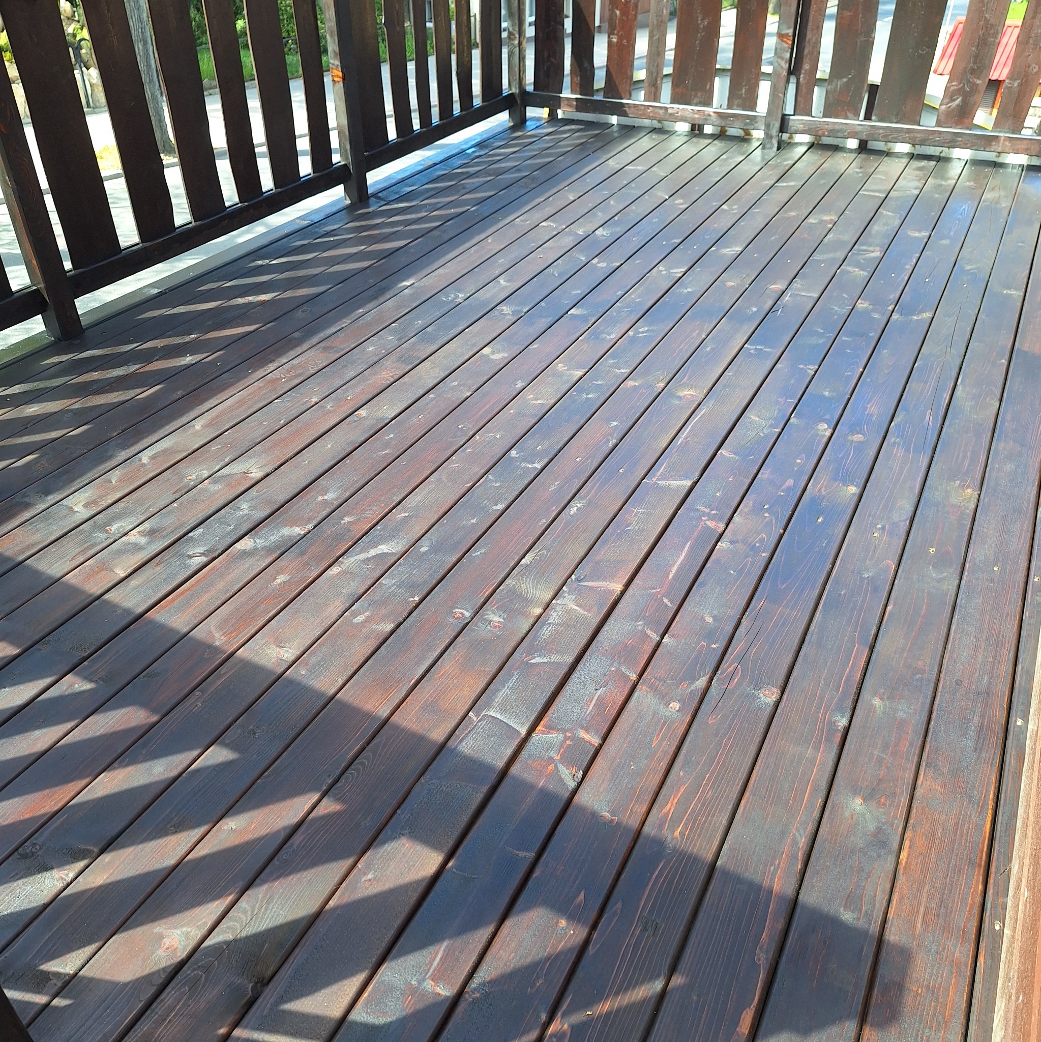 Świeżo polakierowany taras z ciemnego drewna z widocznymi refleksami światła słonecznego i cieniem balustrady.