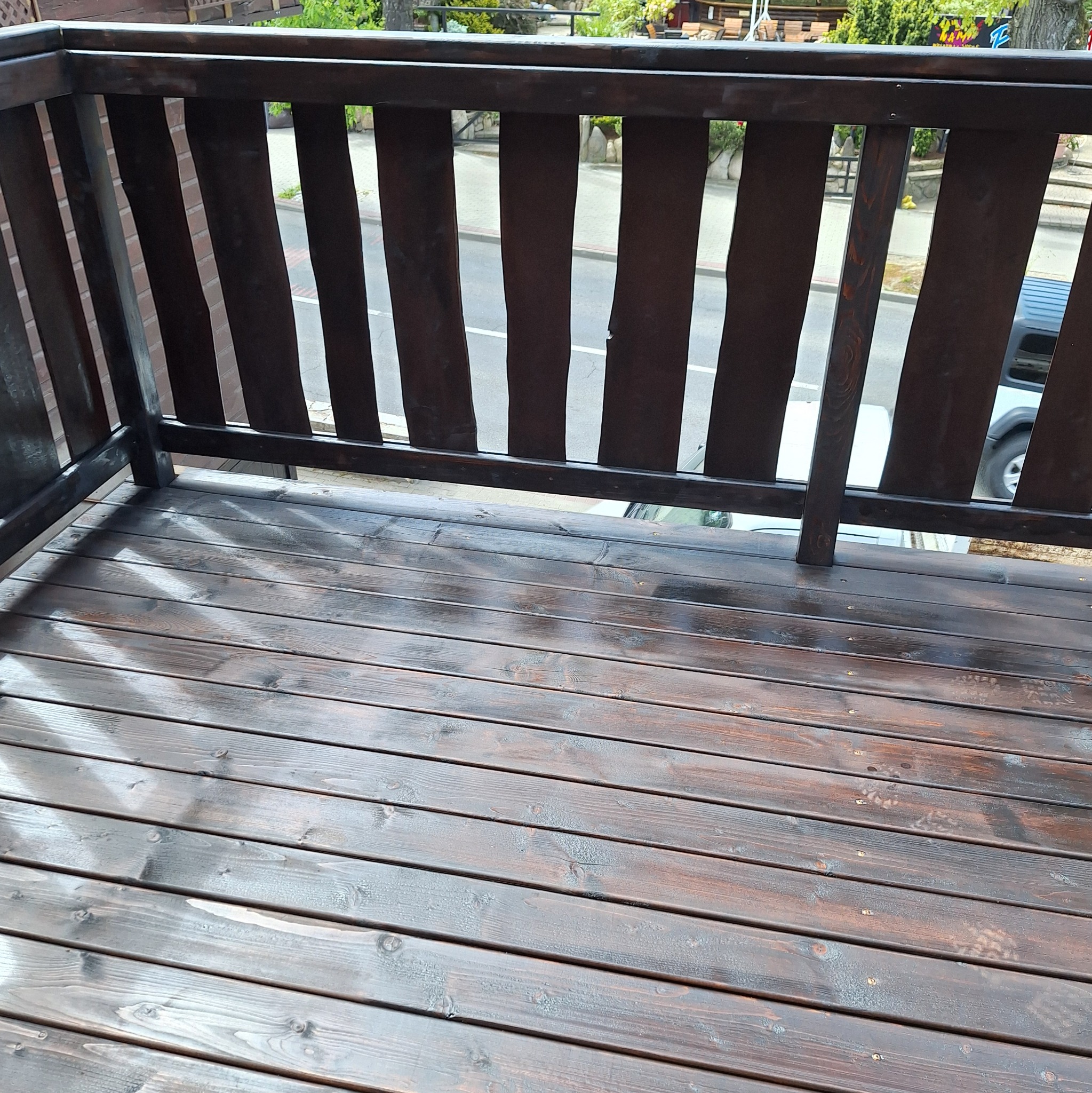 Ciemnobrązowy drewniany taras z balustradą, widok z góry na ulicę i zaparkowany samochód.