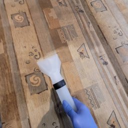 A-Z Cleaning - Czyszczenie tapicerki kanapy urządzeniem ekstrakcyjnym, widoczna ręka w niebieskiej rękawicy i dysza urządzenia na wzorzystym materiale.