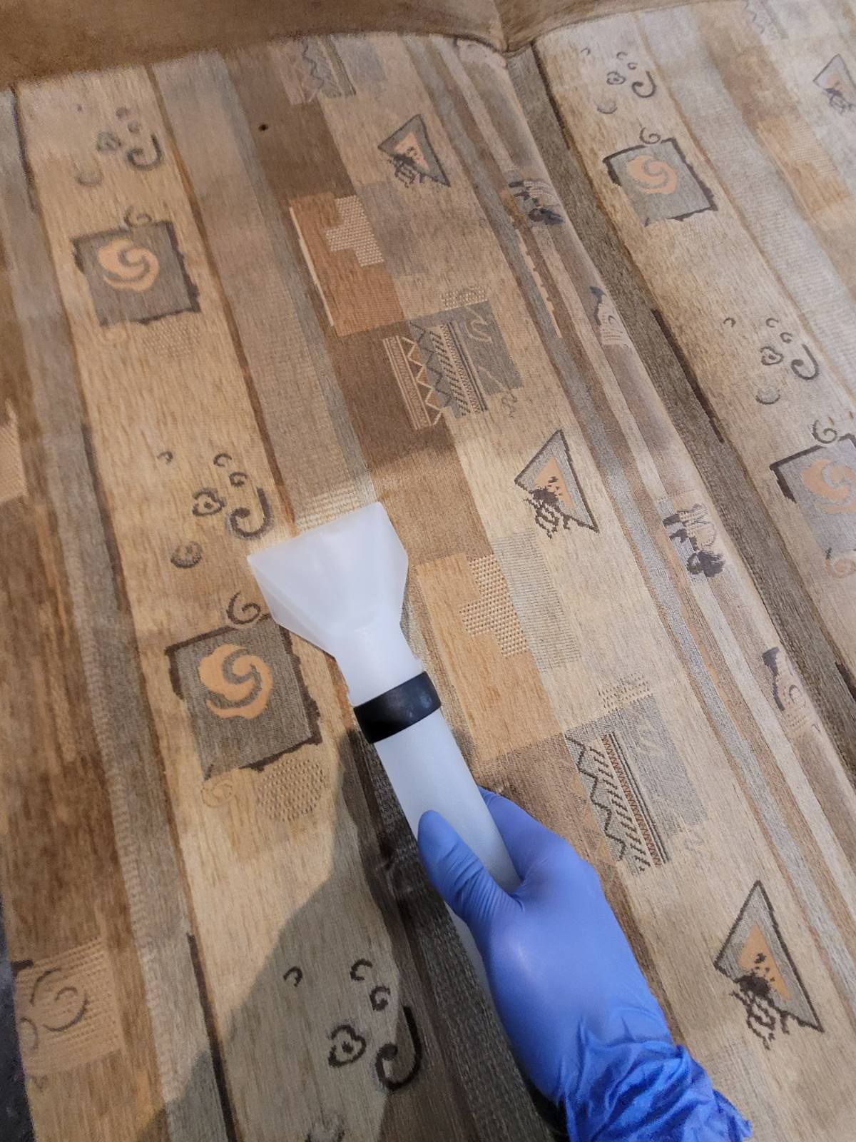 Czyszczenie tapicerki kanapy urządzeniem ekstrakcyjnym, widoczna ręka w niebieskiej rękawicy i dysza urządzenia na wzorzystym materiale.