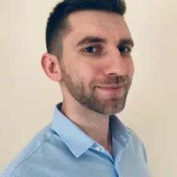 Portret mężczyzny z krótką, ciemną fryzurą i brodą, ubranego w błękitną koszulę w drobną kratkę, pozującego na tle jednolitej, jasnej ściany.