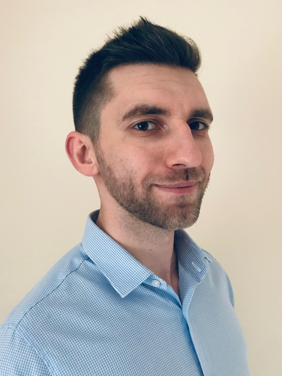 Portret mężczyzny z krótką, ciemną fryzurą i brodą, ubranego w błękitną koszulę w drobną kratkę, pozującego na tle jednolitej, jasnej ściany.