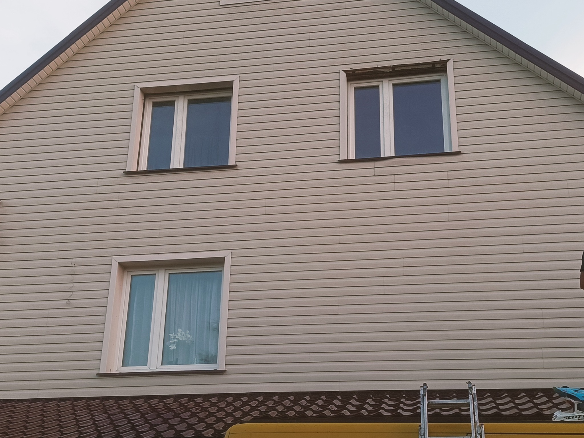 Dwupiętrowy dom z beżową elewacją sidingową, trzema oknami z białymi ramami, widoczny fragment dachu i drabiny, na jednym z okien widoczne ślady po niedokończonym uszczelnianiu.