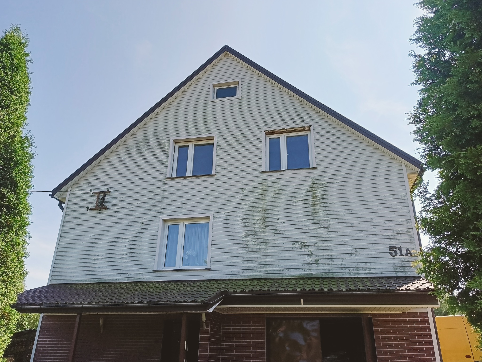 Dwupiętrowy dom z jasną elewacją pokrytą zielonym nalotem, widoczny numer domu 51A, brązowy dach i bujne tuje po bokach.