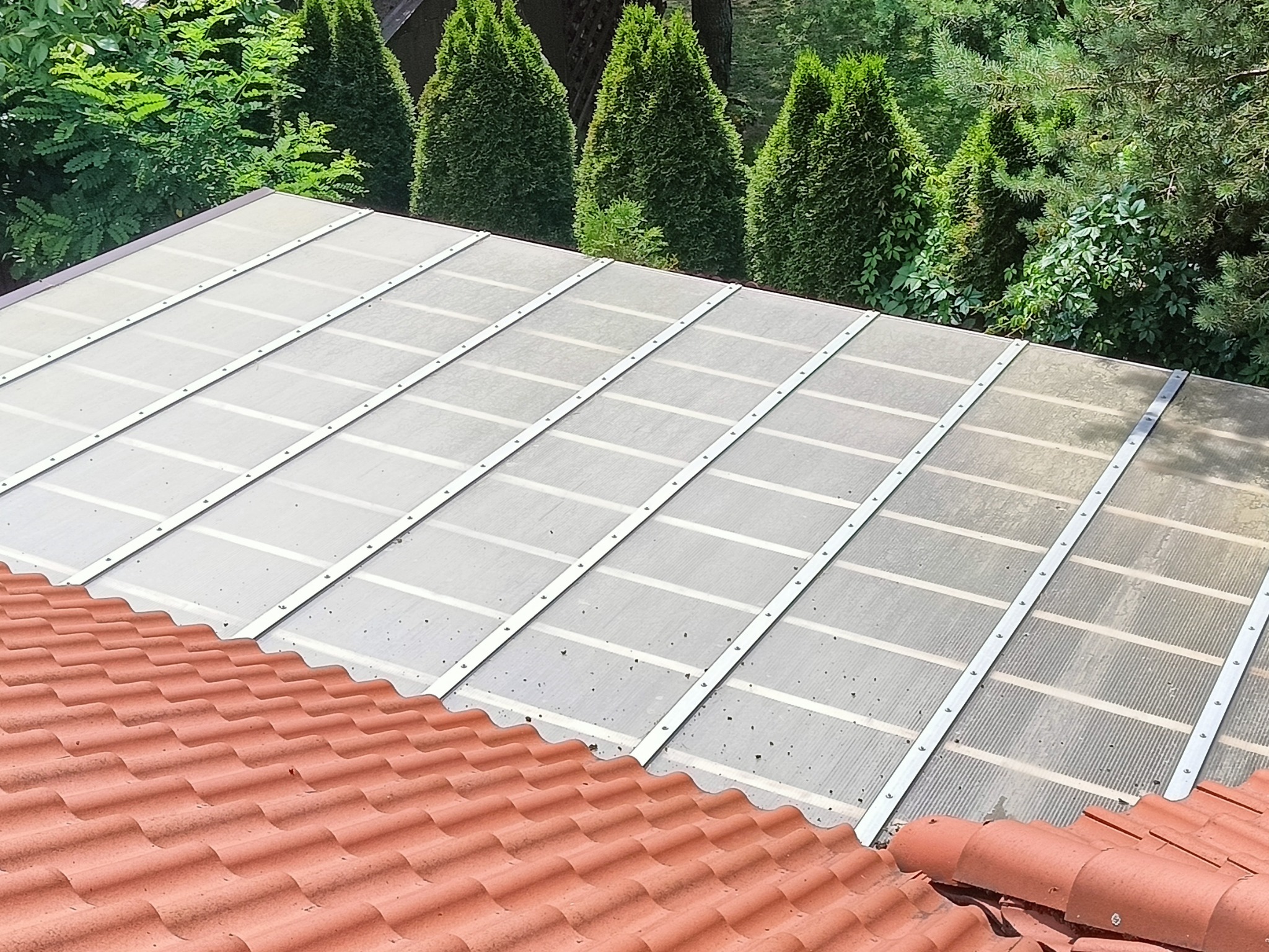 Przezroczyste zadaszenie z poliwęglanu zamontowane nad fragmentem dachu pokrytego czerwoną dachówką, widok z góry na tle zieleni drzew i krzewów.