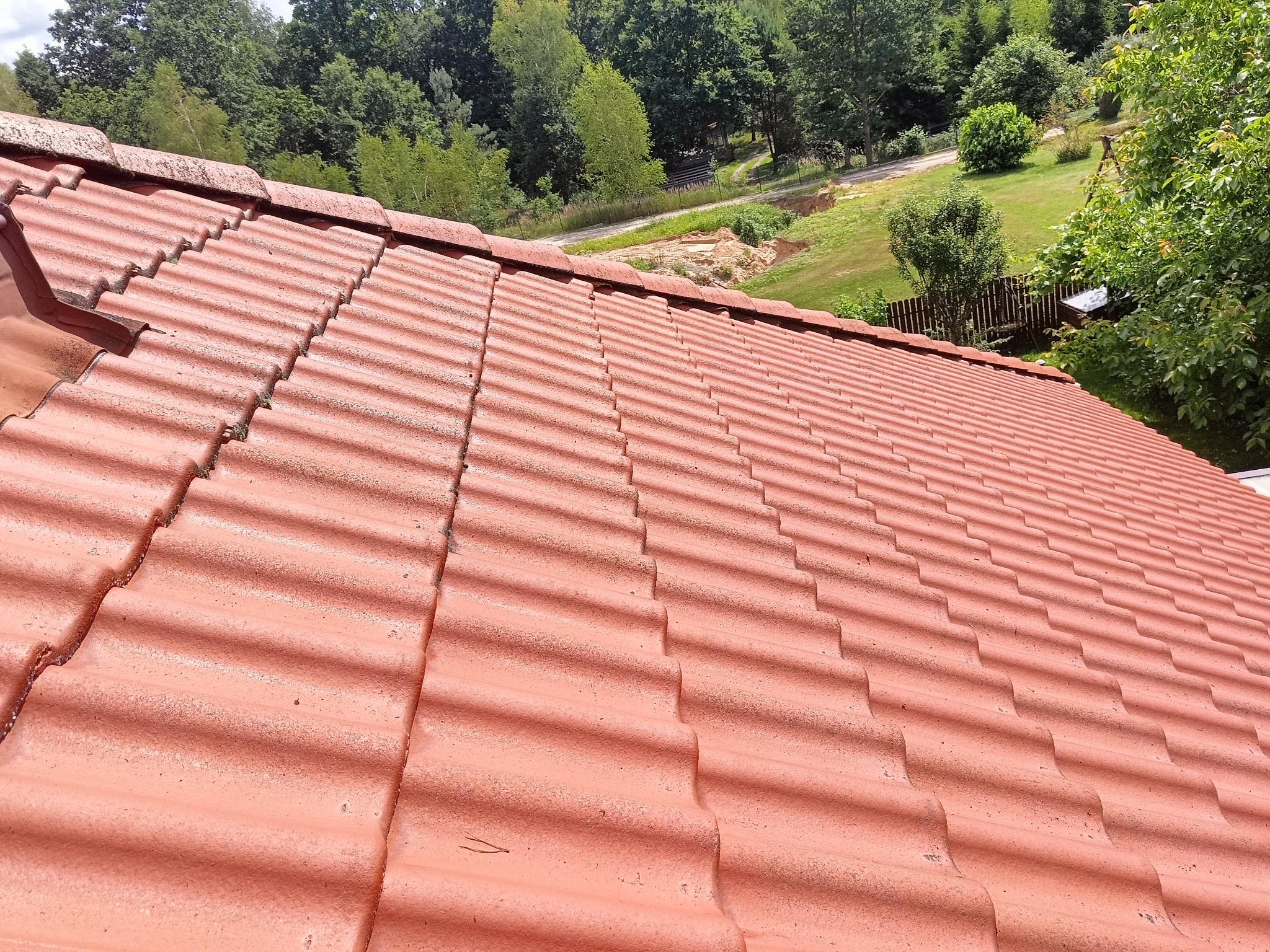 Czerwona dachówka ceramiczna z widocznymi śladami zabrudzeń, z tłem zieleni drzew i krzewów w oddali.