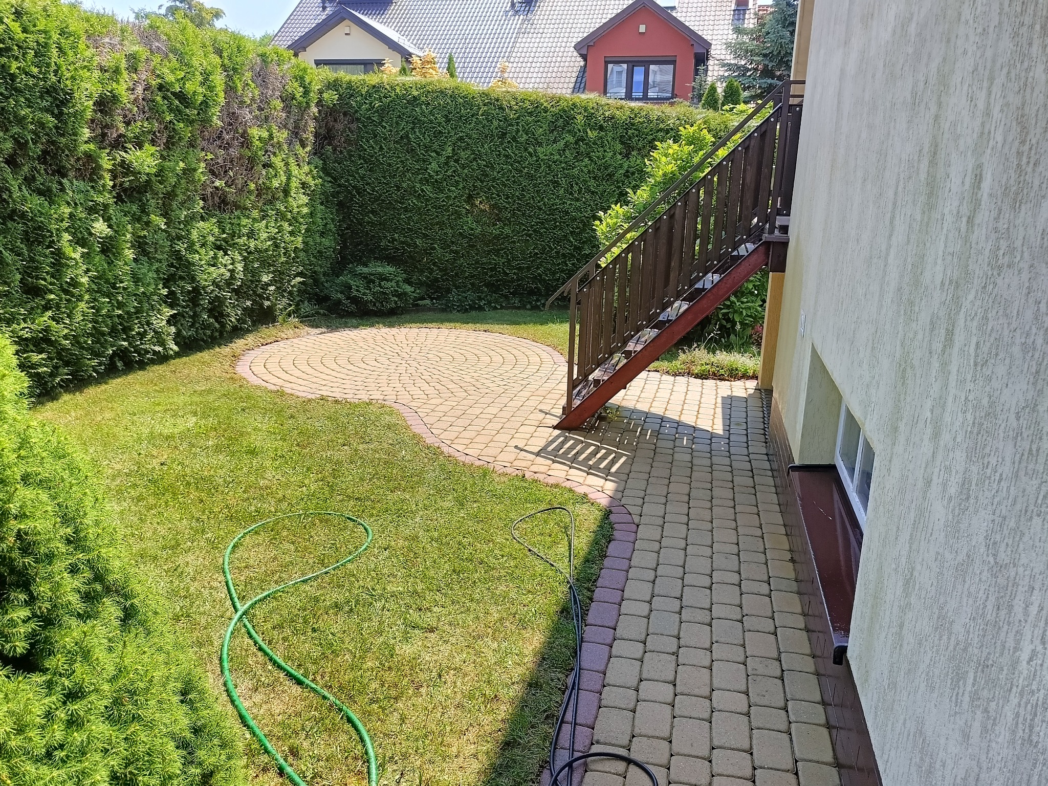 Ułożona z kostki brukowej okrągła powierzchnia w ogrodzie, obok trawnik z zielonym wężem ogrodowym, przy budynku z zewnętrznymi schodami.