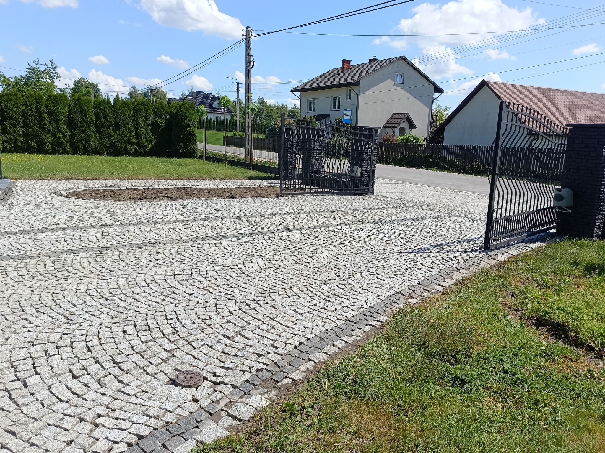 Ułożona kostka brukowa w kształcie wachlarza na podjeździe z otwartą, ozdobną bramą wjazdową. Widoczny trawnik, domy w tle oraz fragment klombu bez roślin.