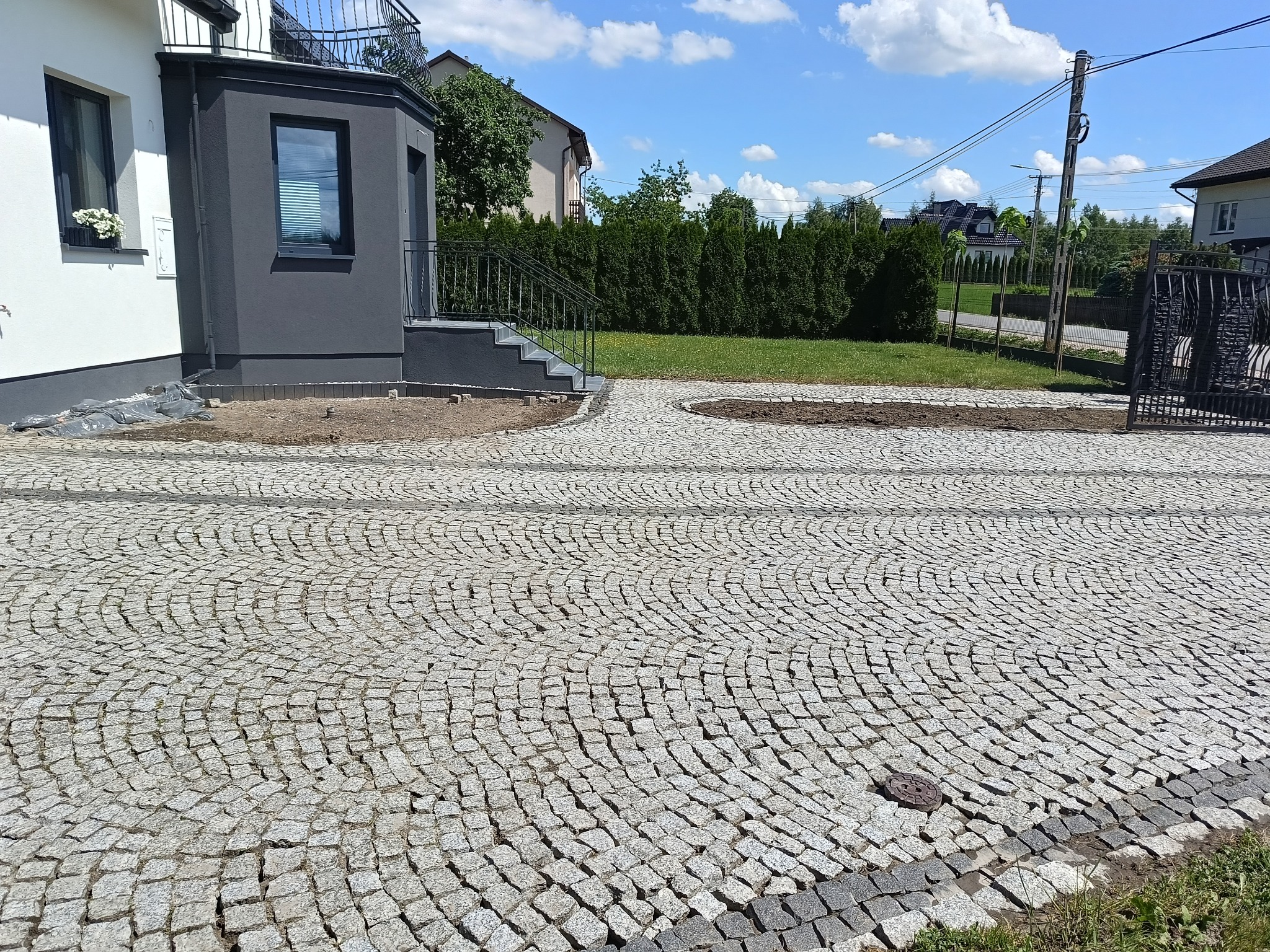 Ułożona z kostki brukowej podjazd do domu z elementami zieleni i świeżo posadzonej trawy, widok na elewację budynku w słoneczny dzień.